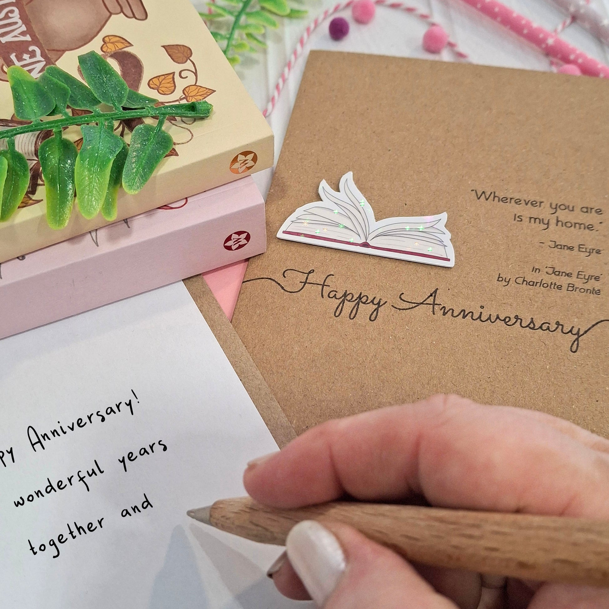 A close up of a hand holding a wooden pen, and writing a happy anniversary message inside a card. The card features a raised holographic embellishment of an open book with gently fanned pages, the words Happy Anniversary, and a quote from the book Jane Eyre by Charlotte Bronte.