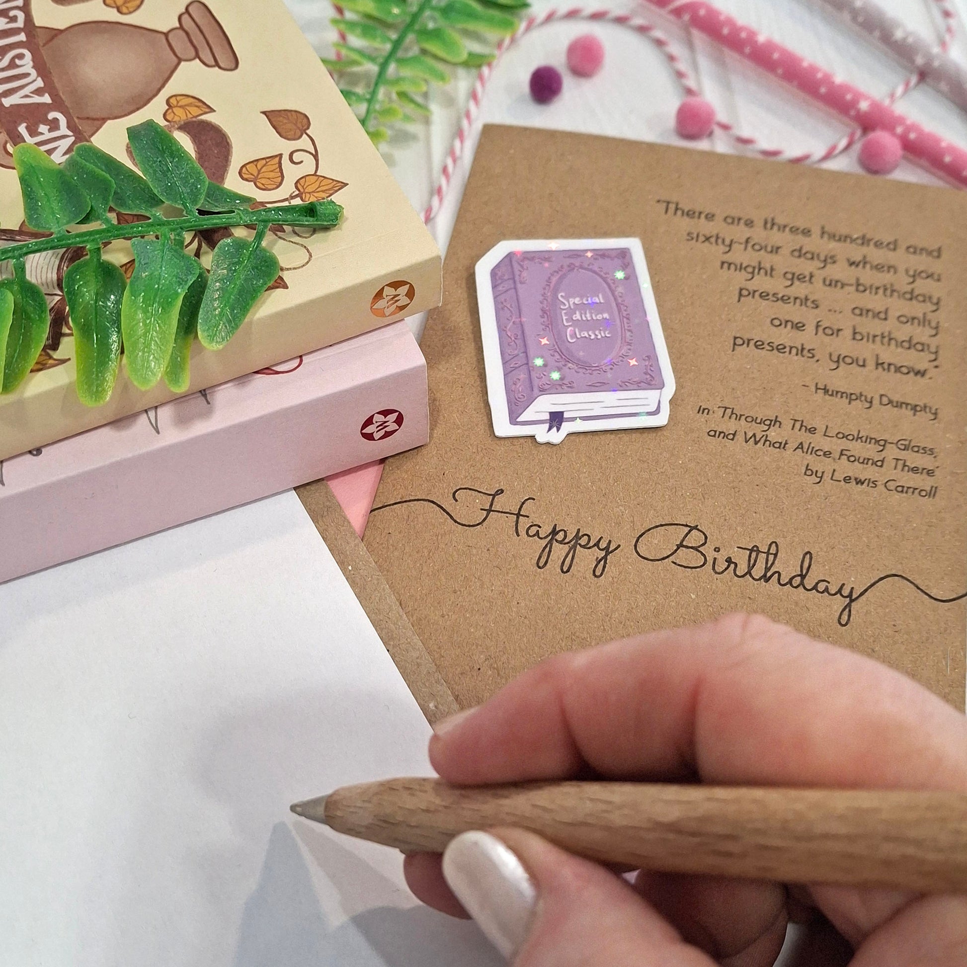 A close-up of a hand holding a wooden pen and writing inside a card. The card features a raised holographic embellishment of a purple special edition classic book, the words Happy Birthday, and a quote from Alice Through the Looking Glass by Lewis Carroll.