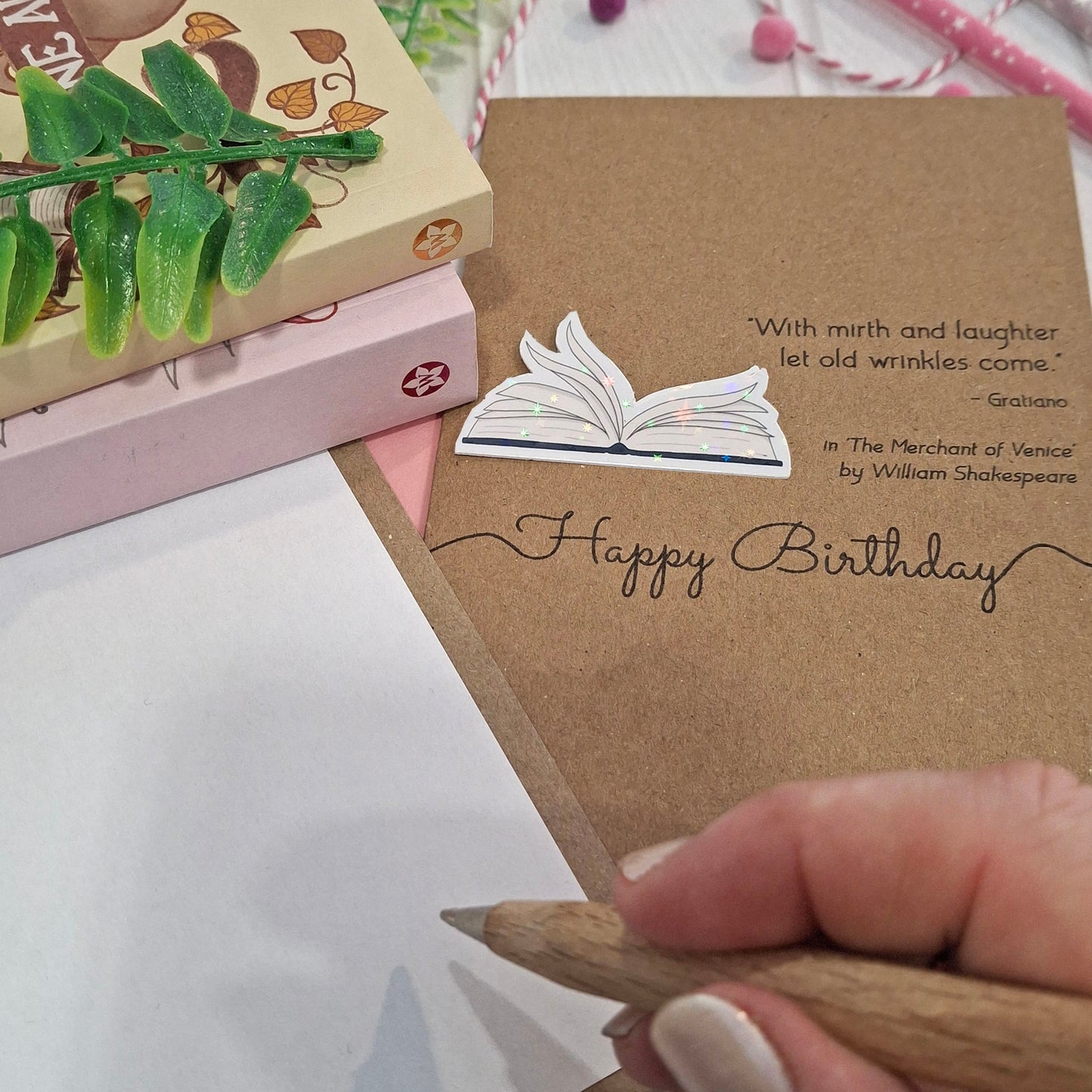 A close up of a hand holding a wooden pen, and writing inside a card. The card features a raised holographic embellishment of an open book with gently fanned pages, the words Happy Birthday, and a quote from The Merchant of Venice by William Shakespeare.