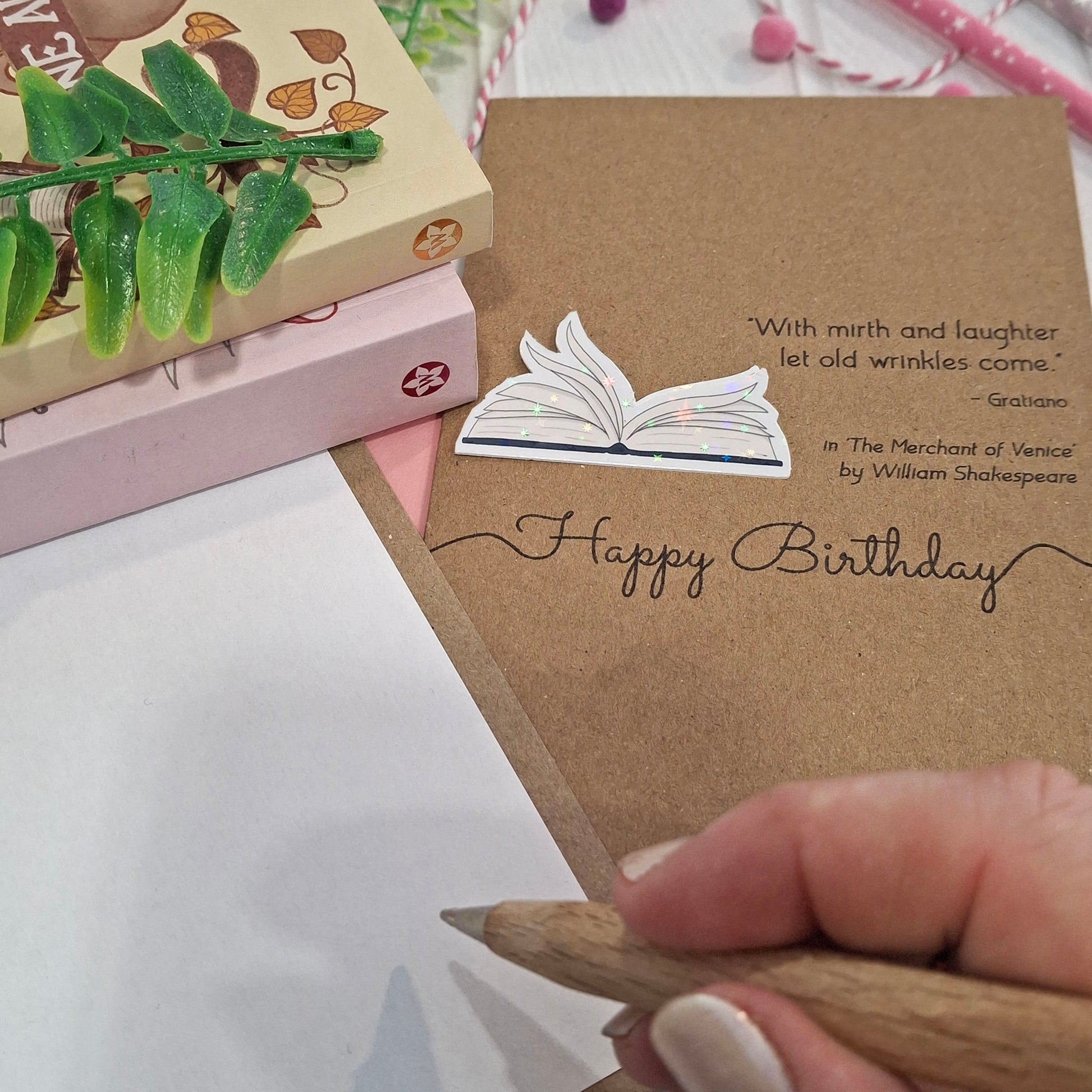 A close up of a hand holding a wooden pen, and writing inside a card. The card features a raised holographic embellishment of an open book with gently fanned pages, the words Happy Birthday, and a quote from The Merchant of Venice by William Shakespeare.