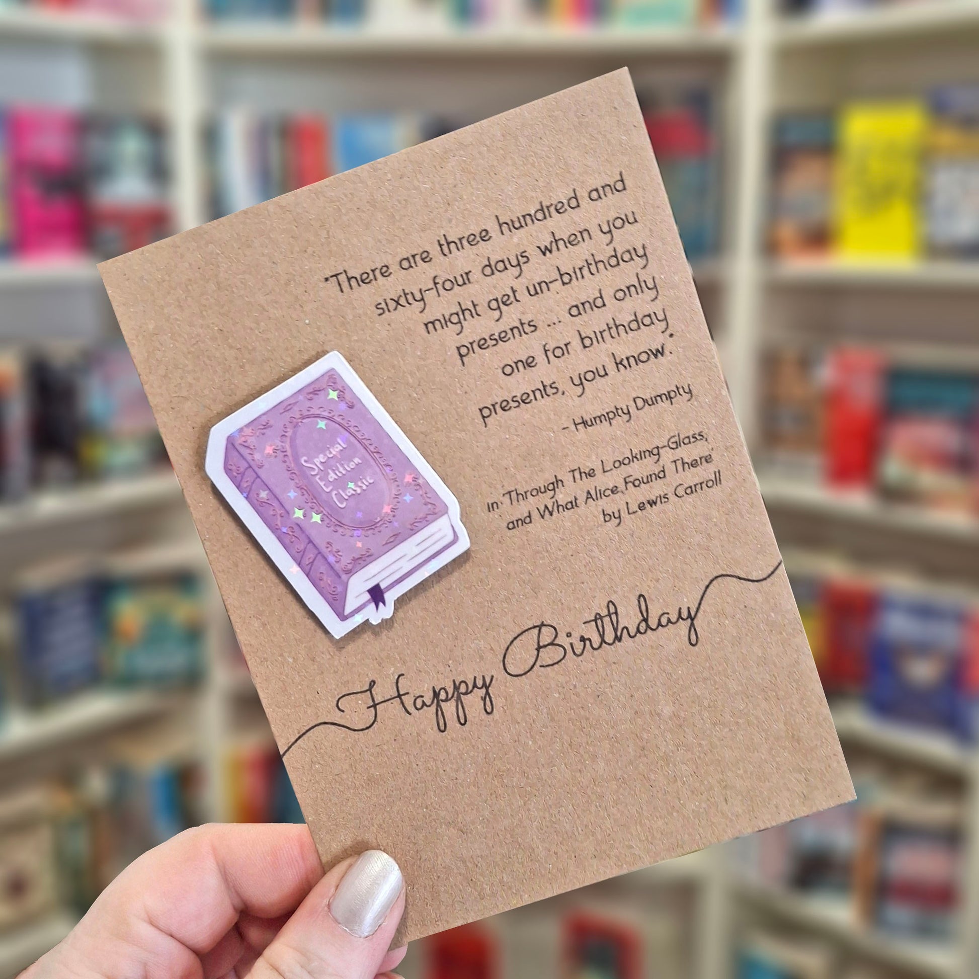 A hand holds a brown kraft happy birthday card featuring a raised holographic embellishment of a purple special edition classic book, and a printed quote by Lewis Carroll. The background shows a blurred bookshop interior.