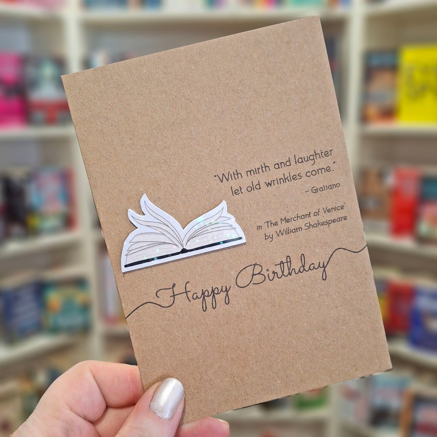 A hand holds a brown kraft happy birthday card featuring a raised holographic embellishment of an open book with gently fanned pages, and a printed quote by William Shakespeare. The background shows a blurred bookshop interior.