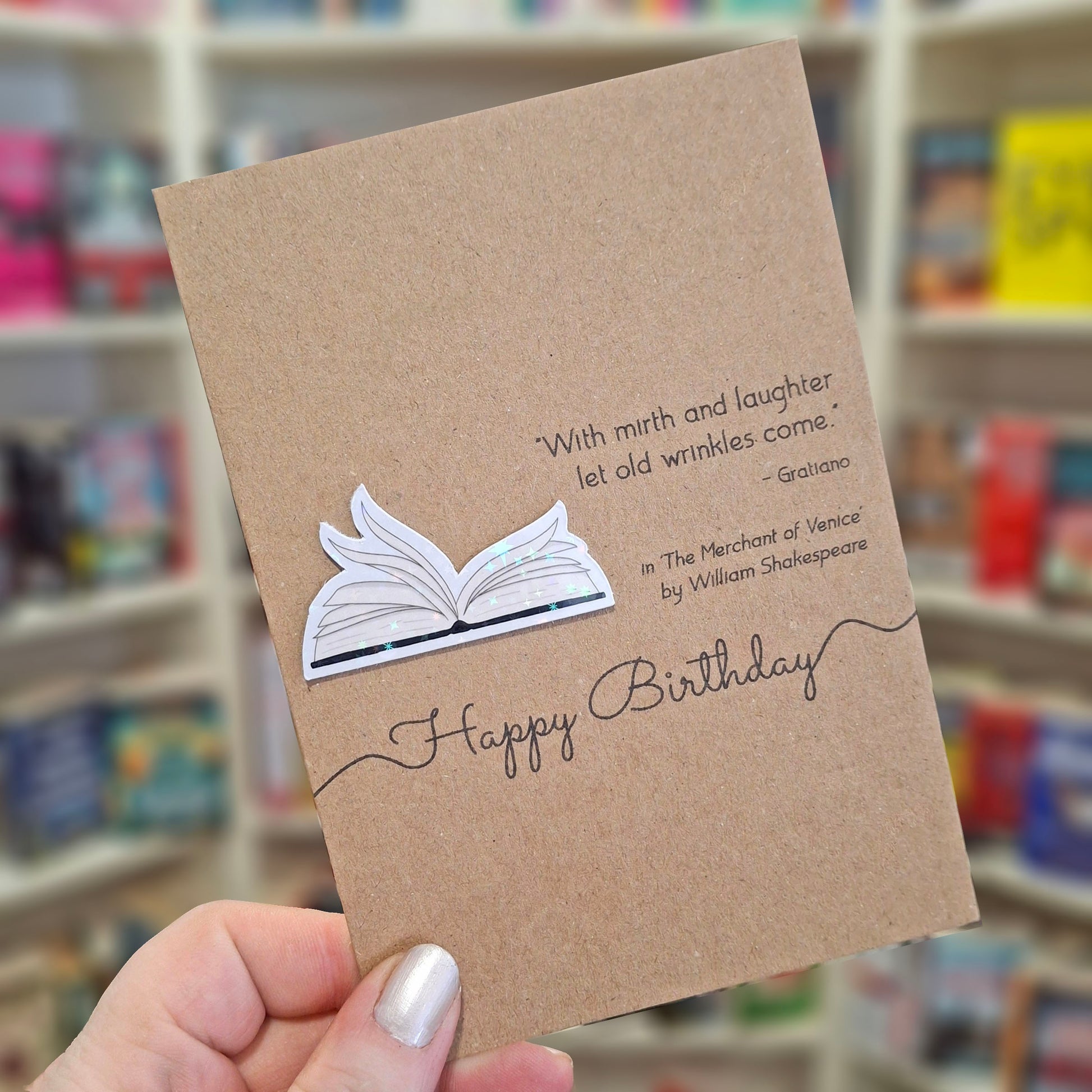 A hand holds a brown kraft happy birthday card featuring a raised holographic embellishment of an open book with gently fanned pages, and a printed quote by William Shakespeare. The background shows a blurred bookshop interior.