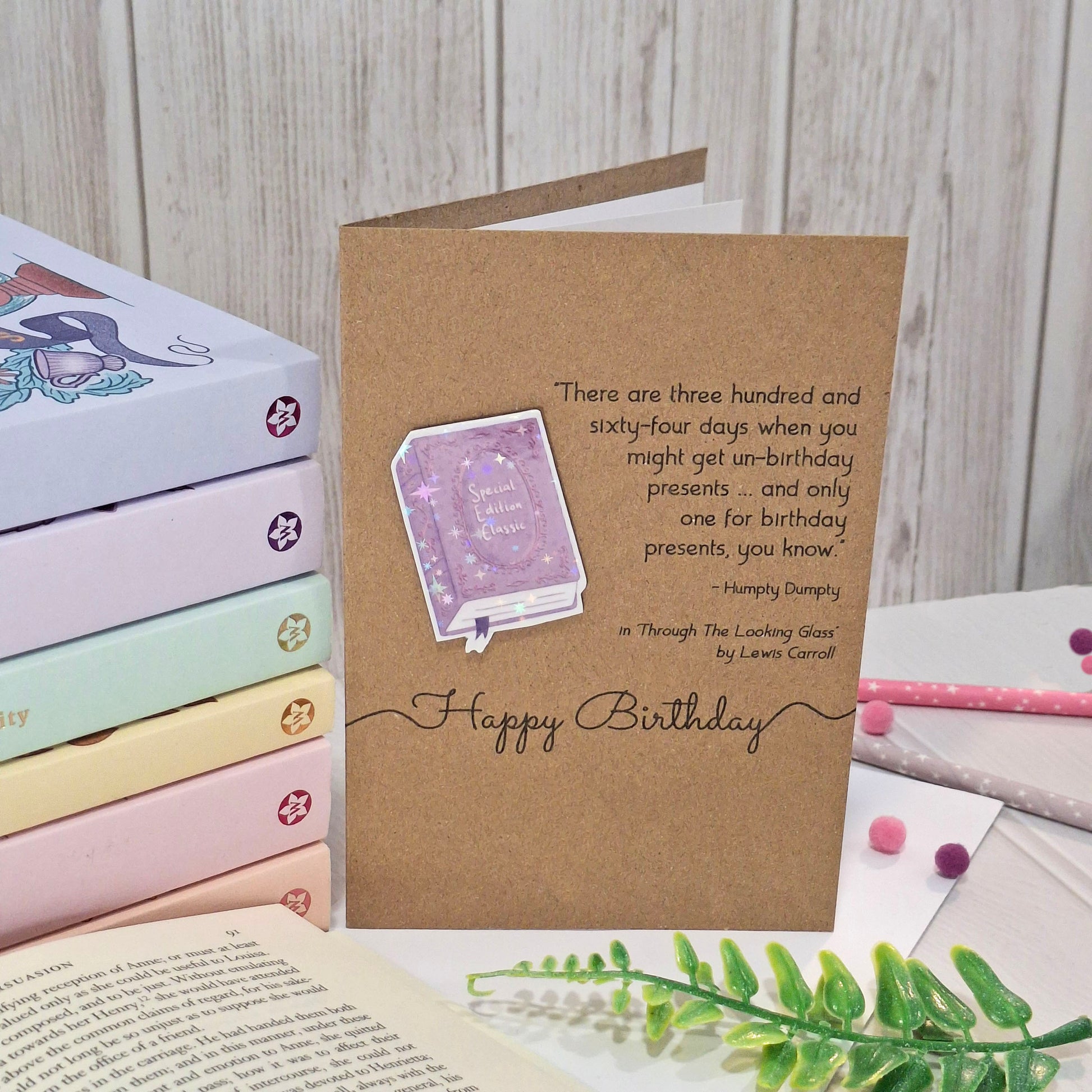 A brown kraft birthday card styled on a tabletop with pastel books, a book open underneath, and green leaves. The card features a raised holographic embellishment of a purple special edition classic book, the words Happy Birthday, and a quote from Alice Through the Looking Glass by Lewis Carroll.
