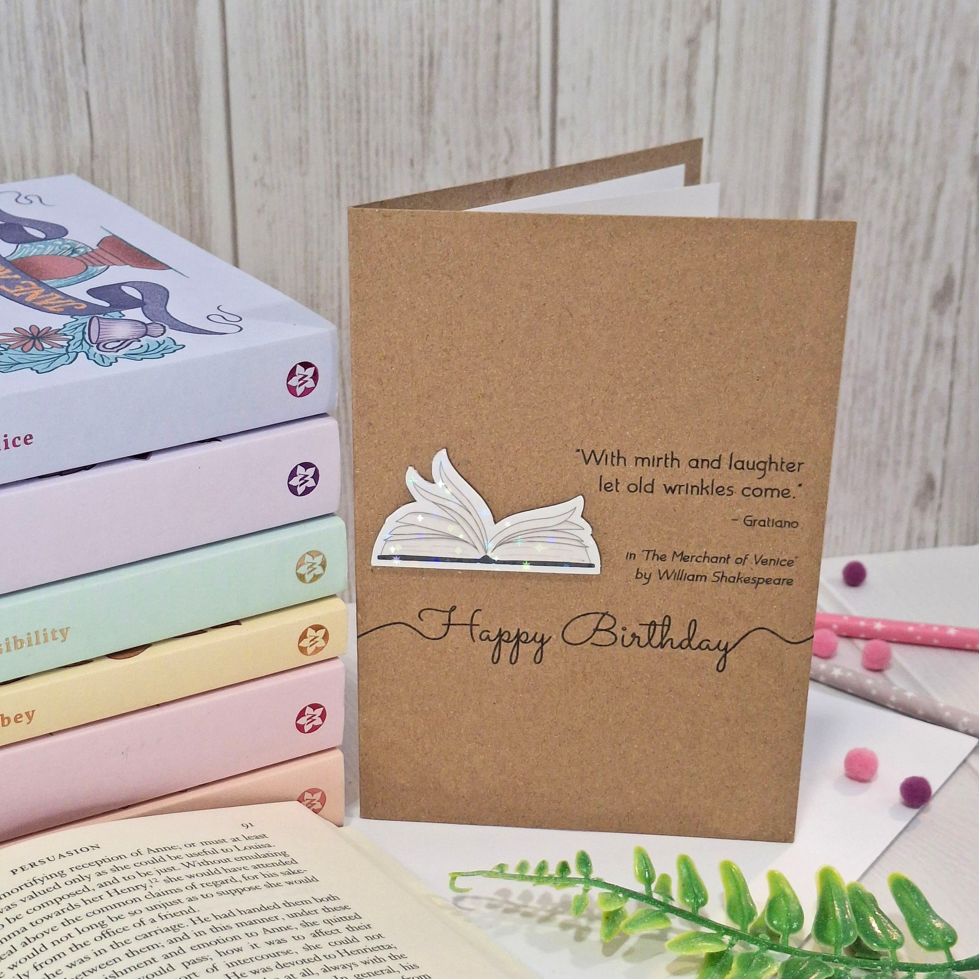 A handmade brown kraft birthday card styled on a white surface with books and a green fern in the background. The card features a raised holographic embellishment of an open book with gently fanned pages, the words Happy Birthday, and a quote from The Merchant of Venice by William Shakespeare.