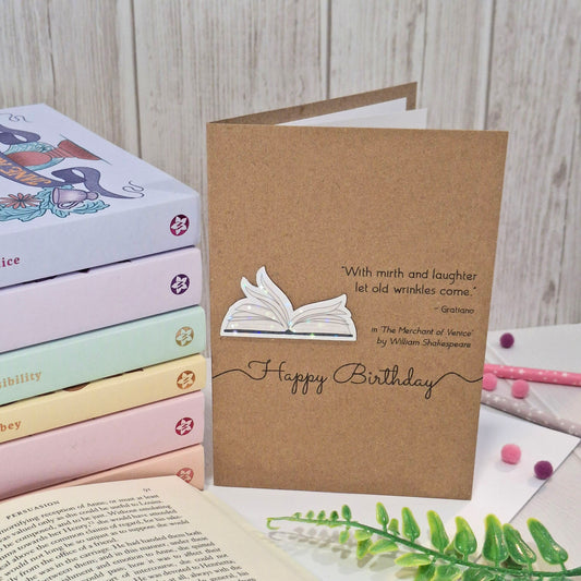A handmade brown kraft birthday card styled on a white surface with books and a green fern in the background. The card features a raised holographic embellishment of an open book with gently fanned pages, the words Happy Birthday, and a quote from The Merchant of Venice by William Shakespeare.