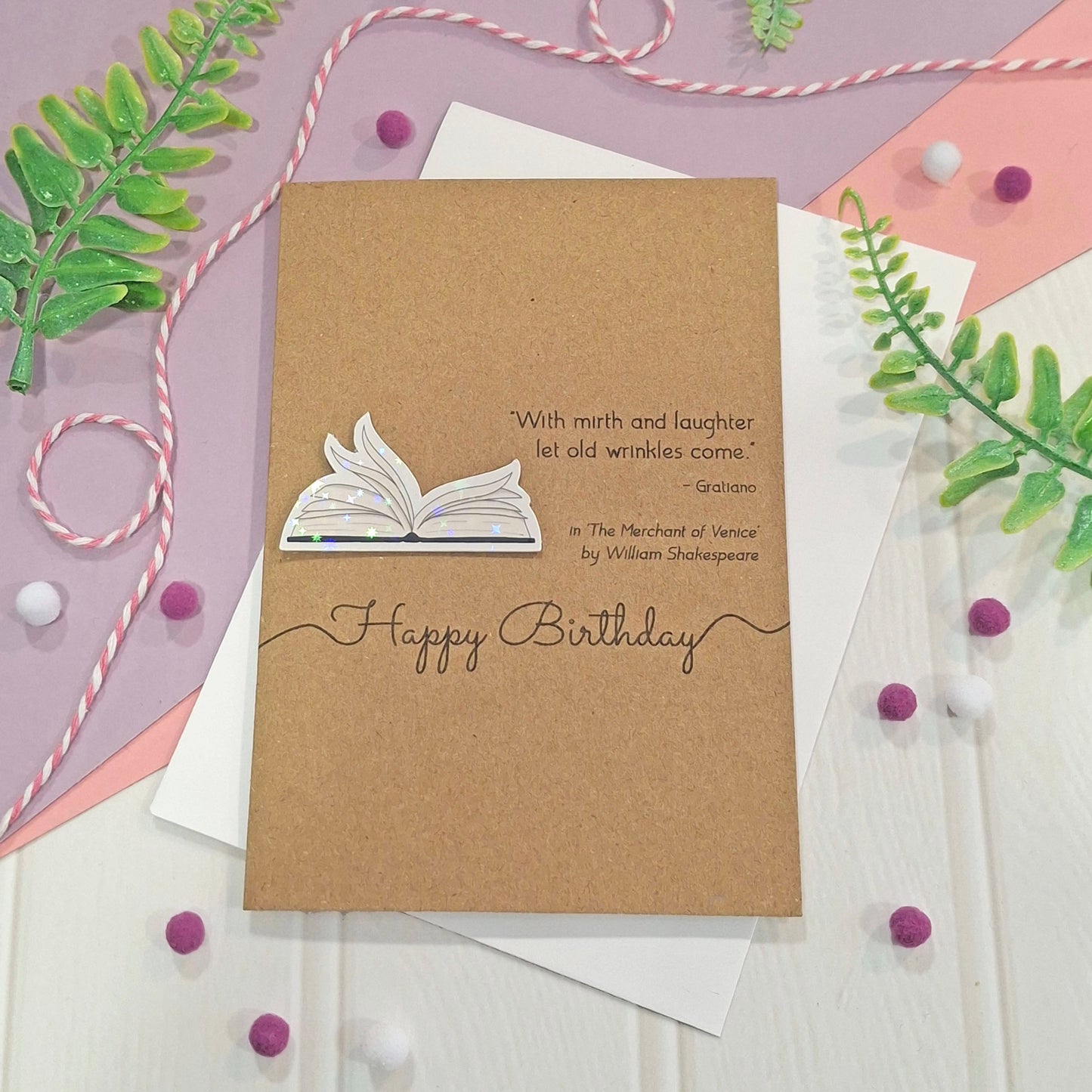 A handmade brown kraft birthday card with illustrated book pages in a raised, holographic embellishment, the words 'Happy Birthday', and a Shakespeare quote, all displayed on a white and pink flat-lay background with decorative green leaves and beads.