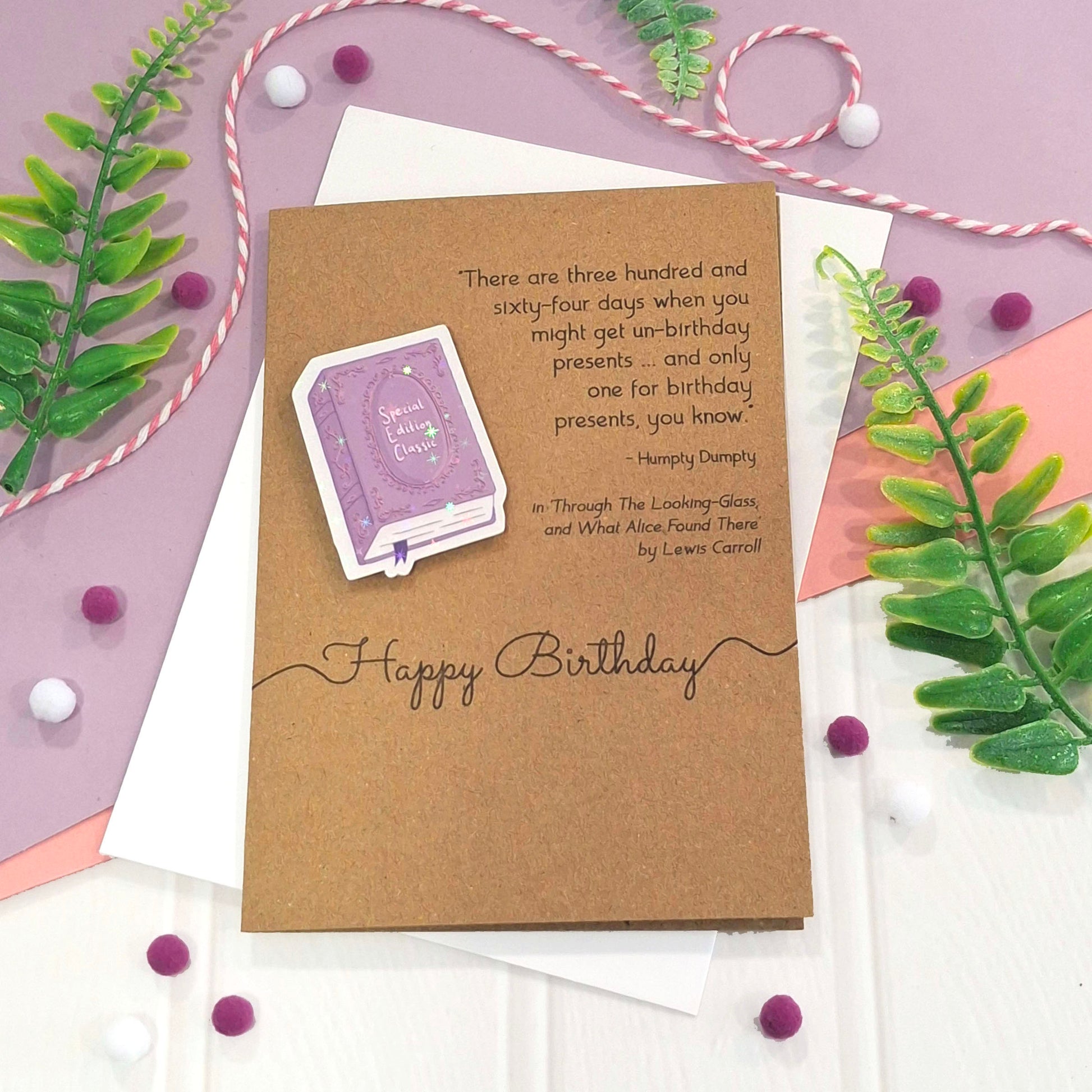 A kraft birthday card with a raised, holographic, illustrated story book embellishment in purple, a quote from Through the Looking Glass by Lewis Carroll, and the words 'Happy Birthday', styled in a flat lay with green leaves, white and pink twine, and small purple and white pom-poms.
