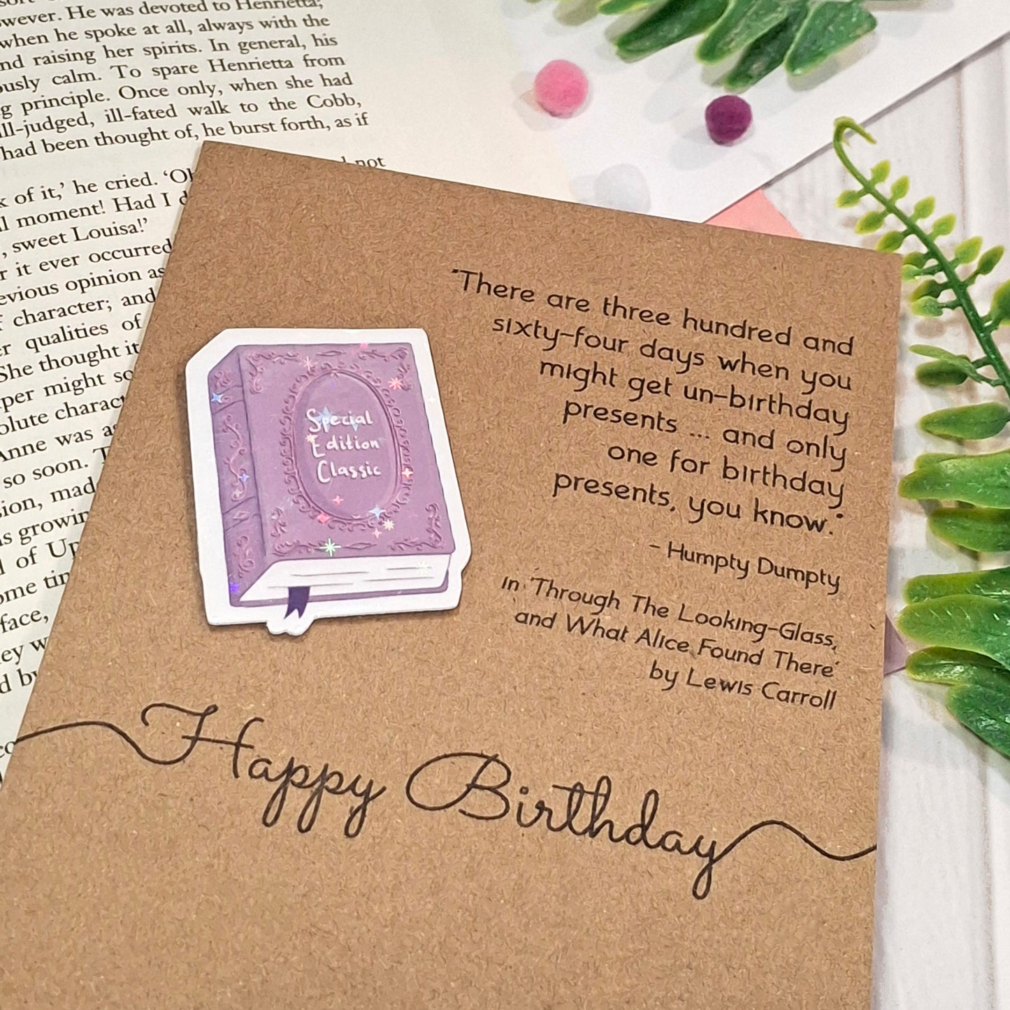 A kraft birthday card styled on top of an open book with green leaves and a pink ribbon. The card features a raised holographic embellishment of a purple special edition classic book, the words Happy Birthday, and a quote from Alice Through the Looking Glass by Lewis Carroll.