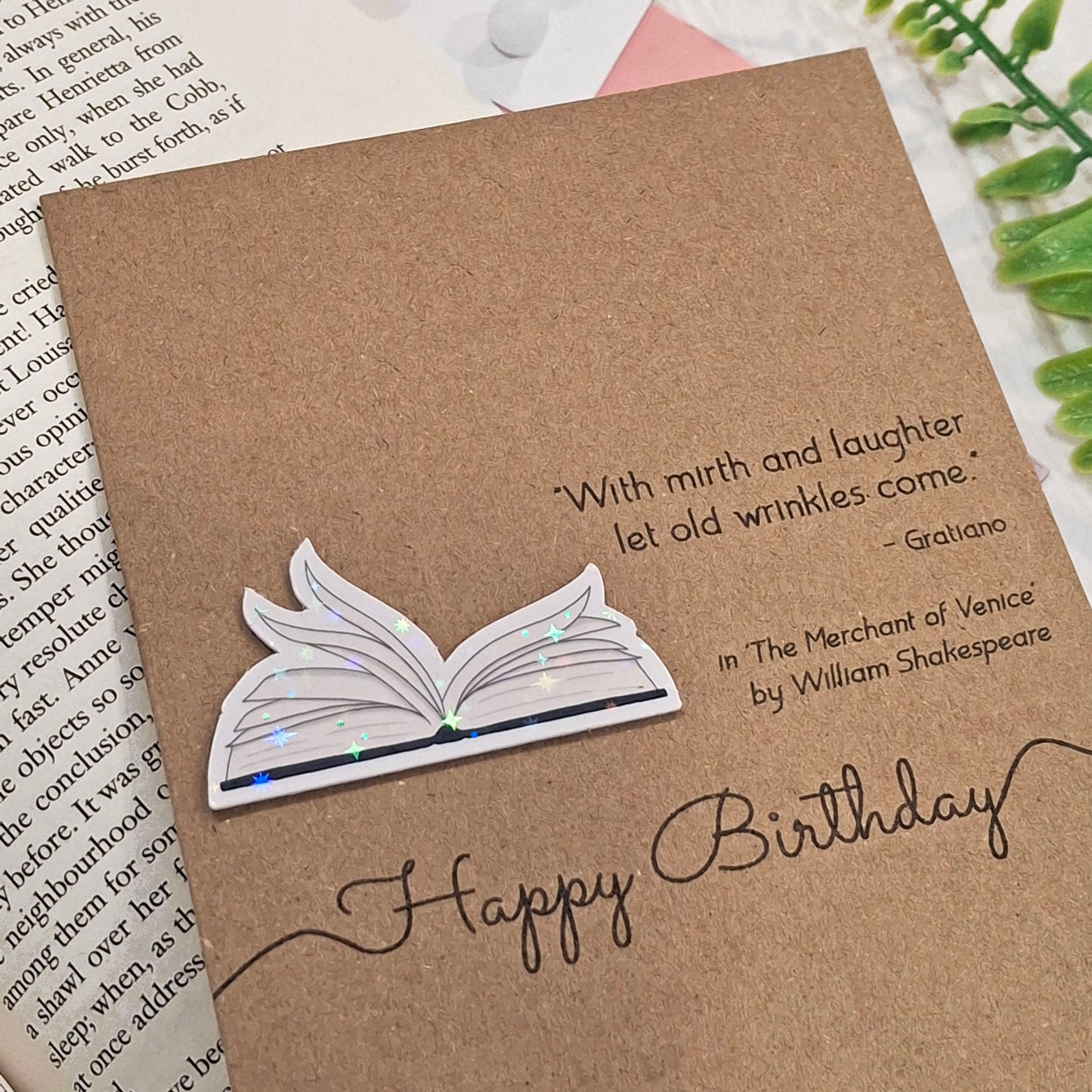 A brown kraft birthday card styled on an open book page with a green fern in the background. The card features a raised holographic embellishment of an open book with gently fanned pages, the words Happy Birthday, and a quote from The Merchant of Venice by William Shakespeare.