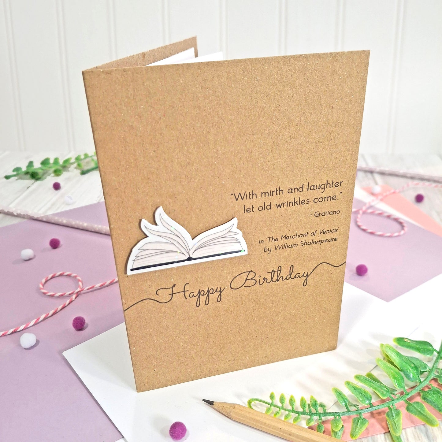 A handmade brown kraft birthday card with illustrated book pages in a raised, holographic embellishment, the words 'Happy Birthday' and a Shakespeare quote, displayed on a styled purple and white flat-lay surface with decorative props.