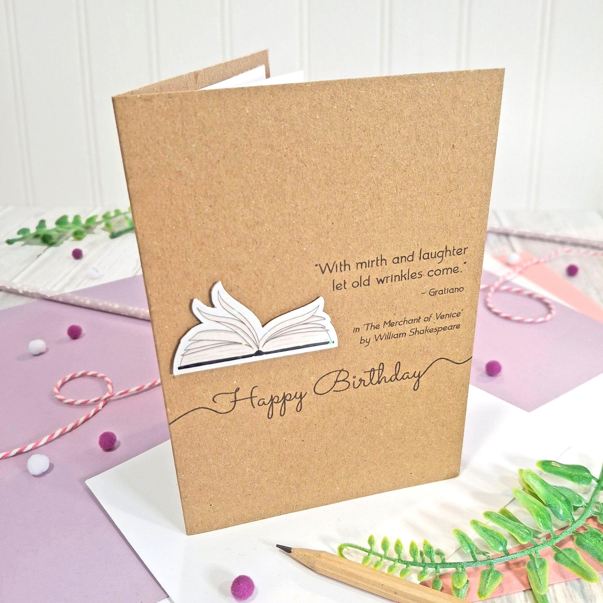 A handmade brown kraft birthday card with illustrated book pages in a raised, holographic embellishment, the words 'Happy Birthday' and a Shakespeare quote, displayed on a styled purple and white flat-lay surface with decorative props.