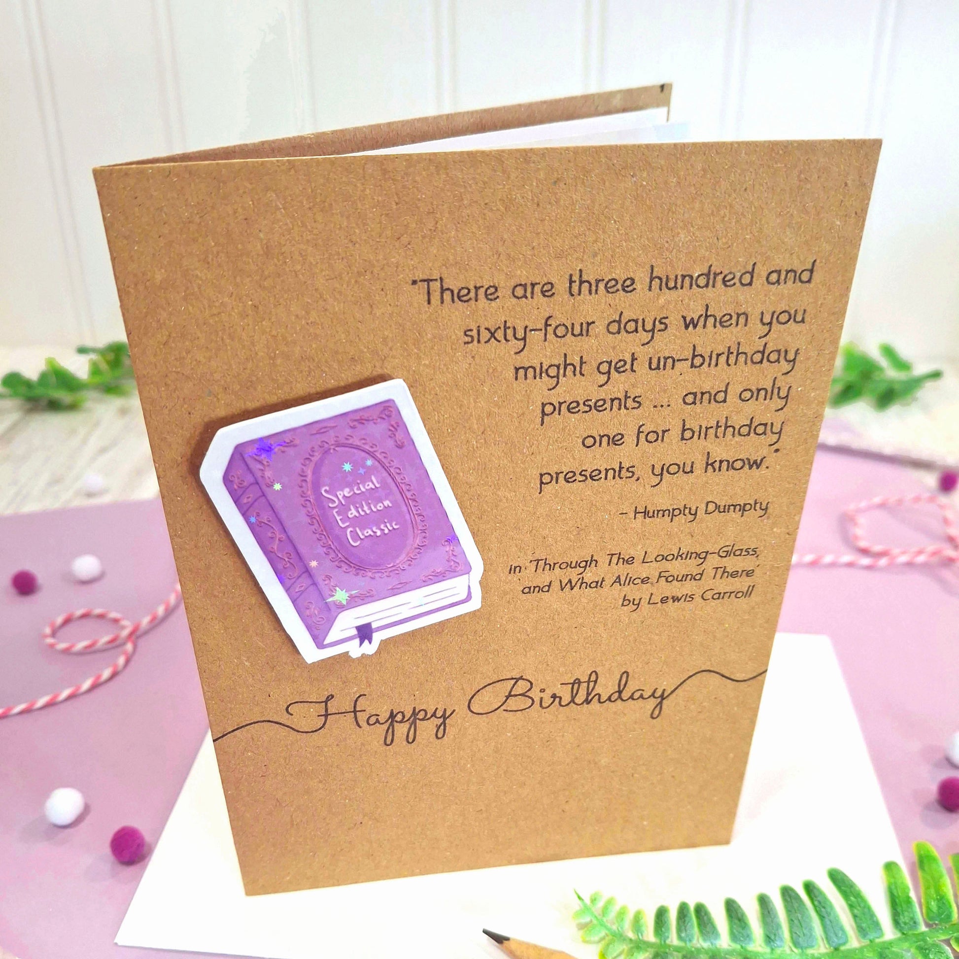 A kraft birthday card with a raised, holographic, illustrated story book embellishment in purple, a quote from Through the Looking Glass by Lewis Carroll, and the words 'Happy Birthday', displayed on a purple surface with decorative props.