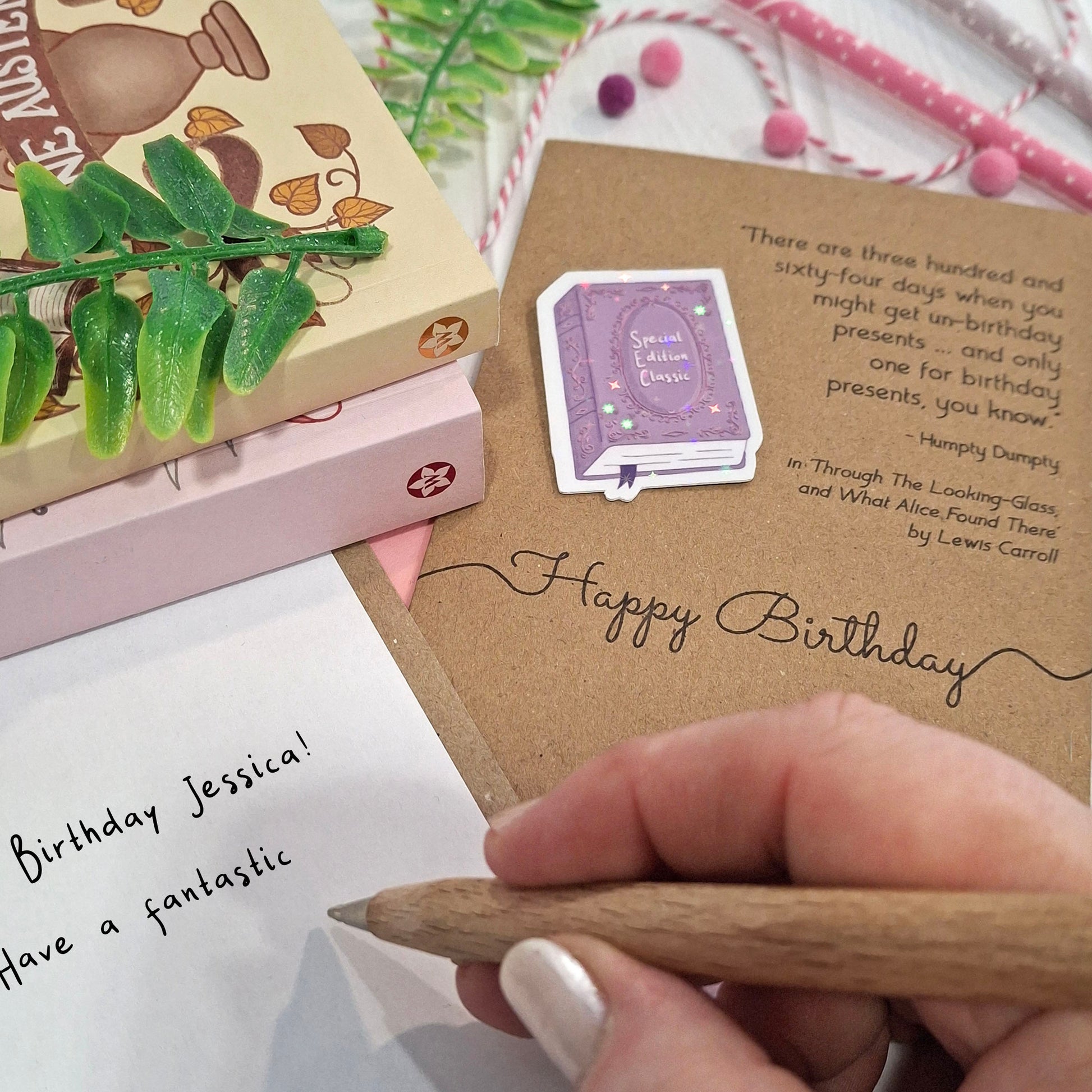 A close-up of a hand holding a wooden pen and writing a happy birthday message inside a card. The card features a raised holographic embellishment of a purple special edition classic book, the words Happy Birthday, and a quote from Alice Through the Looking Glass by Lewis Carroll.