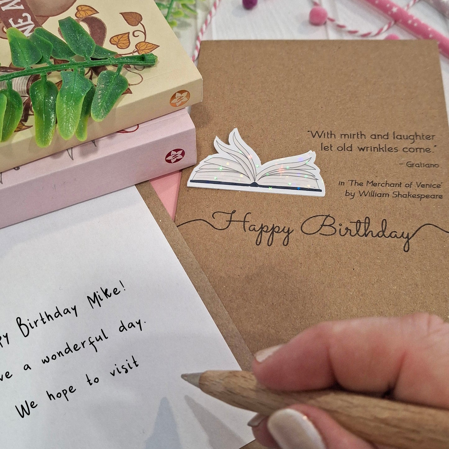 A close up of a hand holding a wooden pen, and writing a happy birthday message inside a card. The card features a raised holographic embellishment of an open book with gently fanned pages, the words Happy Birthday, and a quote from The Merchant of Venice by William Shakespeare.