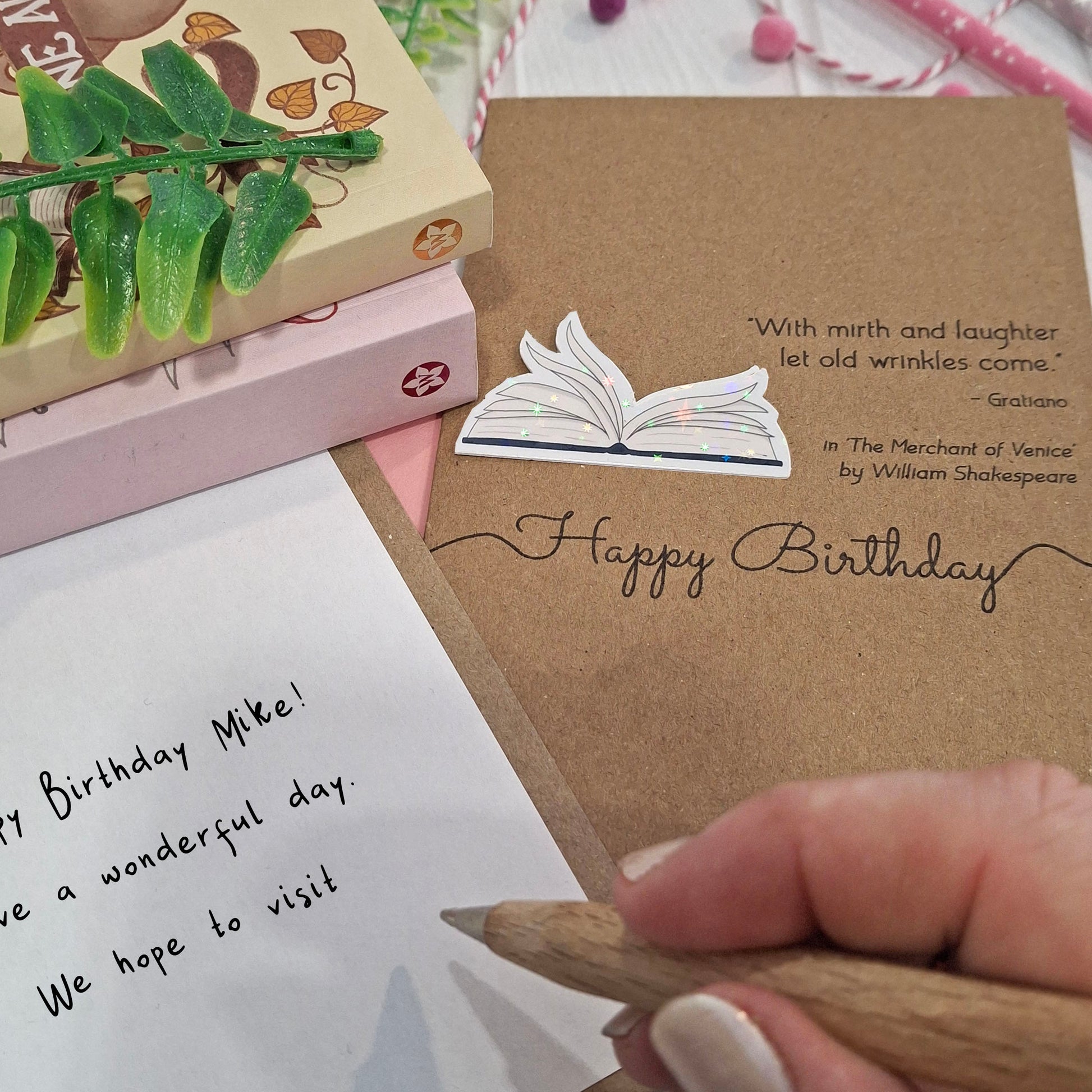 A close up of a hand holding a wooden pen, and writing a happy birthday message inside a card. The card features a raised holographic embellishment of an open book with gently fanned pages, the words Happy Birthday, and a quote from The Merchant of Venice by William Shakespeare.