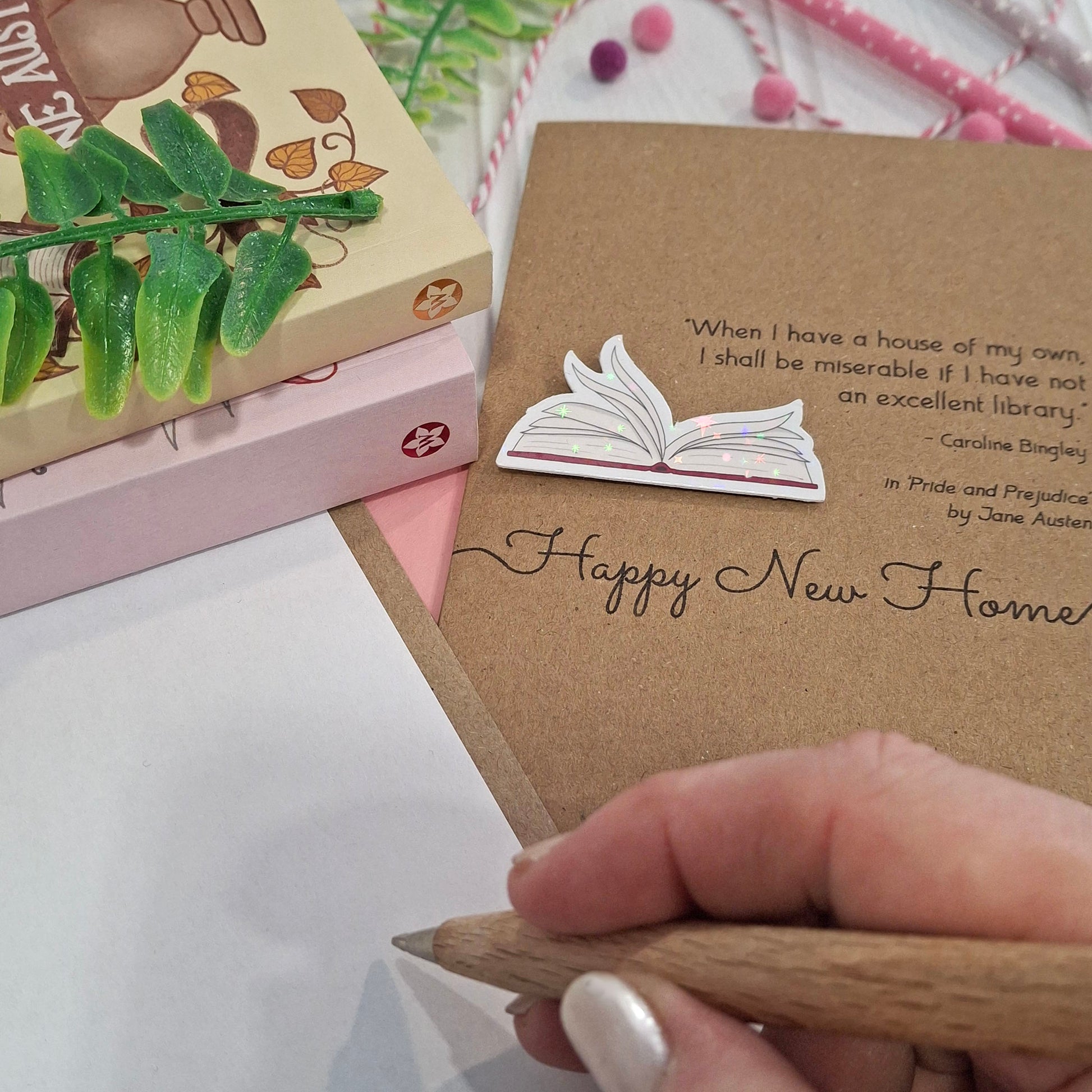A close up of a hand holding a wooden pen, and writing inside a card. The card features a raised holographic embellishment of an open book with gently fanned pages, the words Happy New Home, and a quote from the book Pride and Prejudice by Jane Austen.