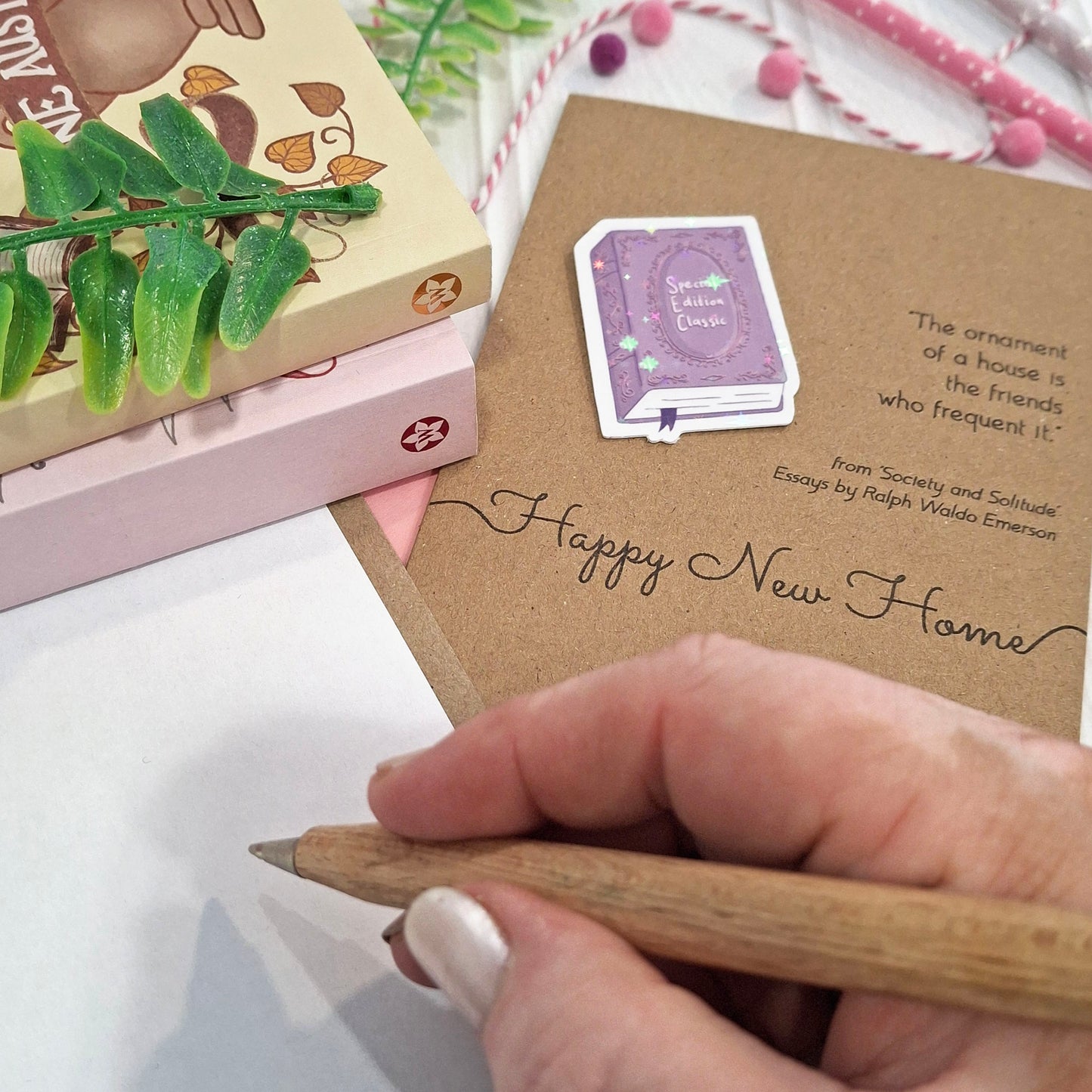 A close-up of a hand holding a wooden pencil writing inside a card. The card features a raised holographic embellishment of a purple special edition classic book, the word Happy New Home, and a quote from Ralph Waldo Emerson.