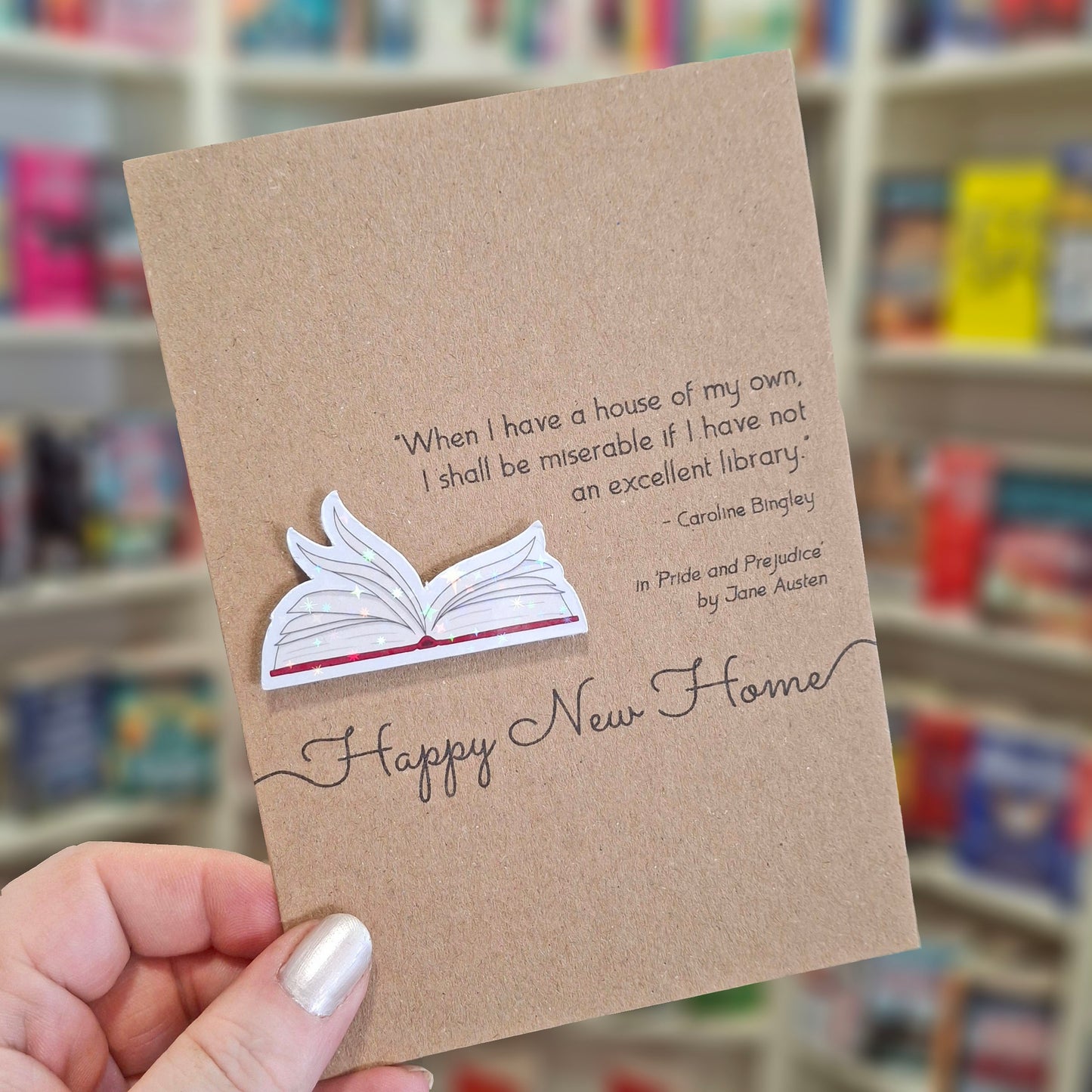 A hand holds a brown kraft happy new home card featuring a raised holographic embellishment of an open book with gently fanned pages, and a printed quote by Jane Austen. The background shows a blurred bookshop interior.