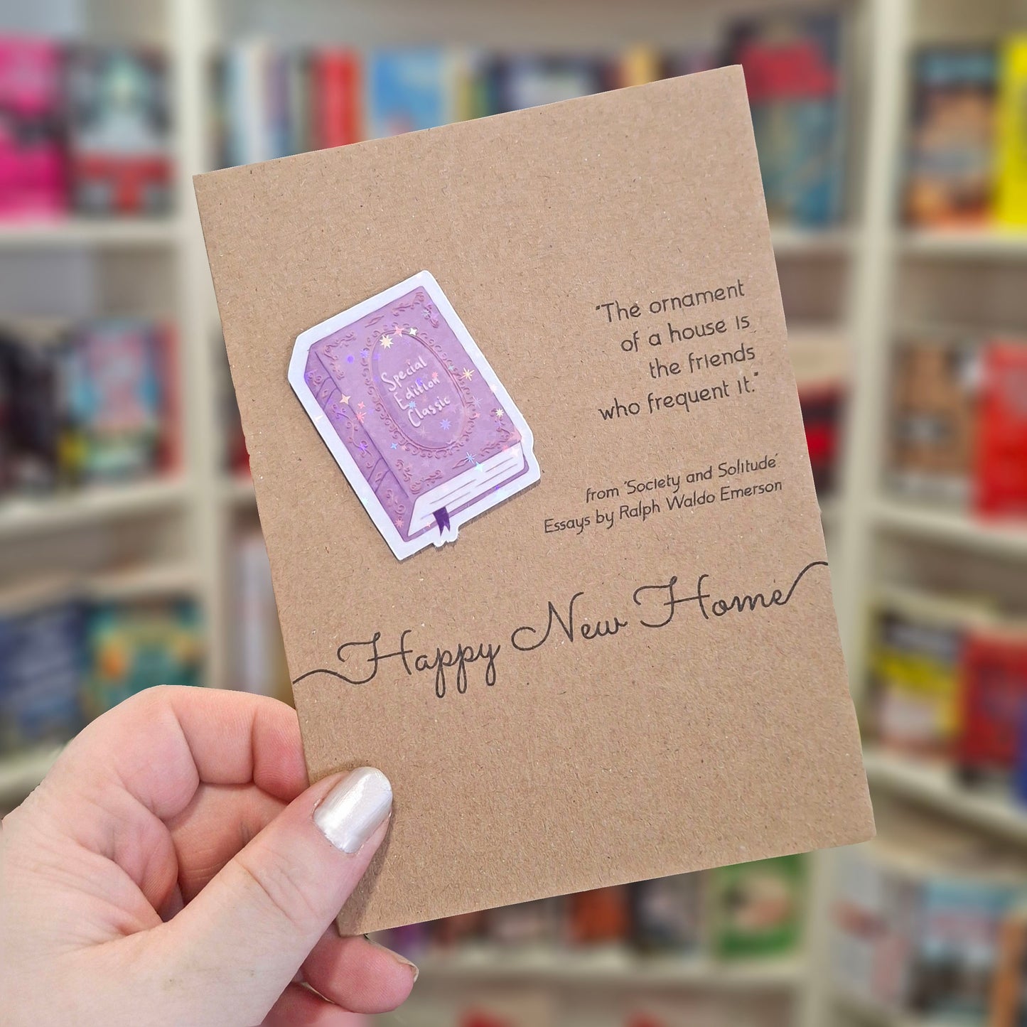 A hand holds a brown kraft happy new home card featuring a raised holographic embellishment of a purple special edition classic book, and a printed quote by Ralph Waldo Emerson. The background shows a blurred bookshop interior.