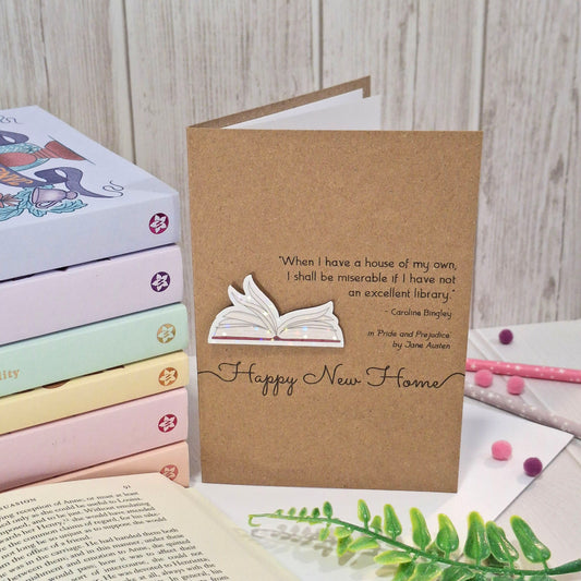 A brown kraft card  styled on a light wood background with books, an open book, and decorative items. The card features a raised holographic embellishment of an open book with gently fanned pages, the words Happy New Home, and a quote from the book Pride and Prejudice by Jane Austen.