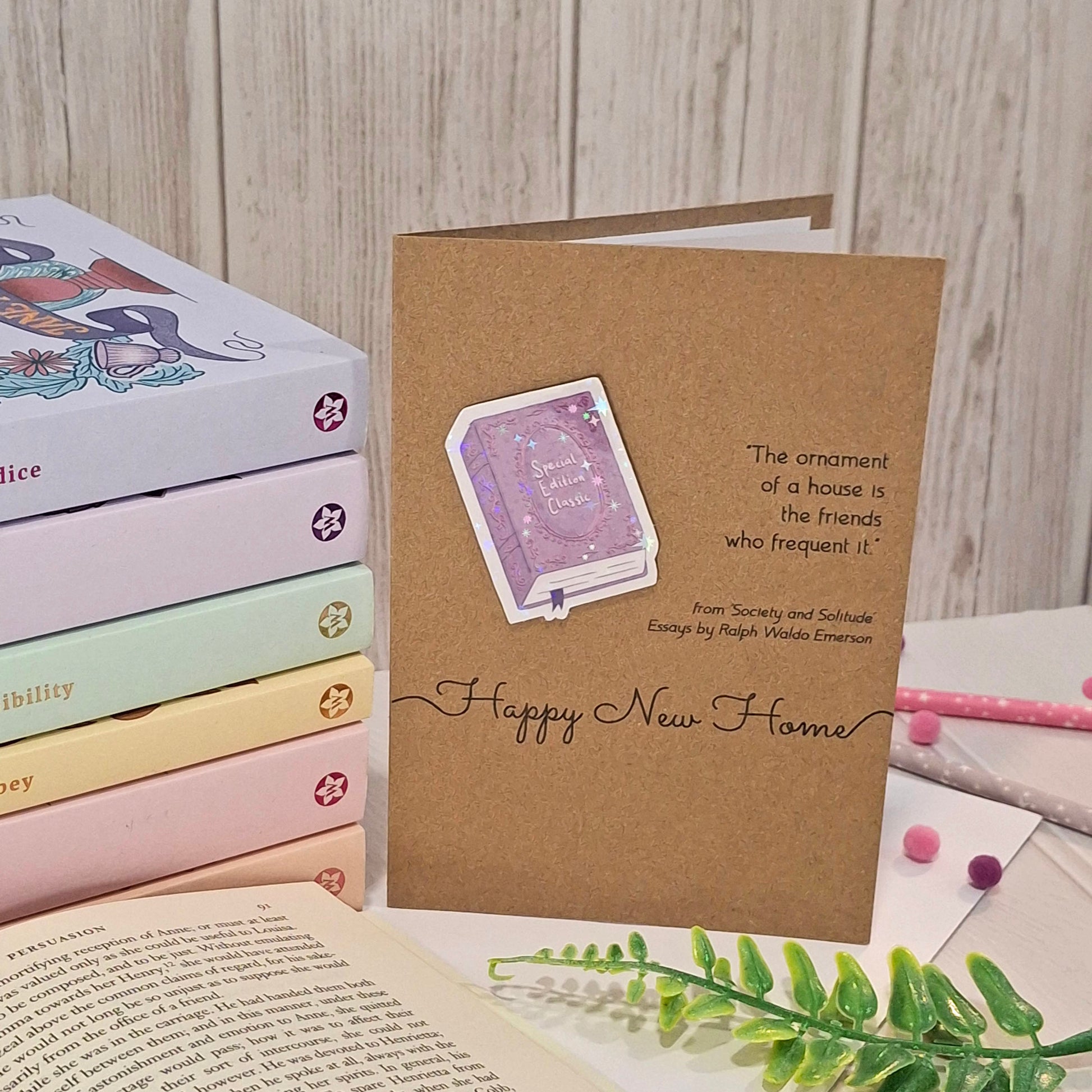 A brown kraft greeting card styled on a tabletop with books, pastel pens, and greenery. The card features a raised holographic embellishment of a purple special edition classic book, the word Happy New Home, and a quote from Ralph Waldo Emerson.
