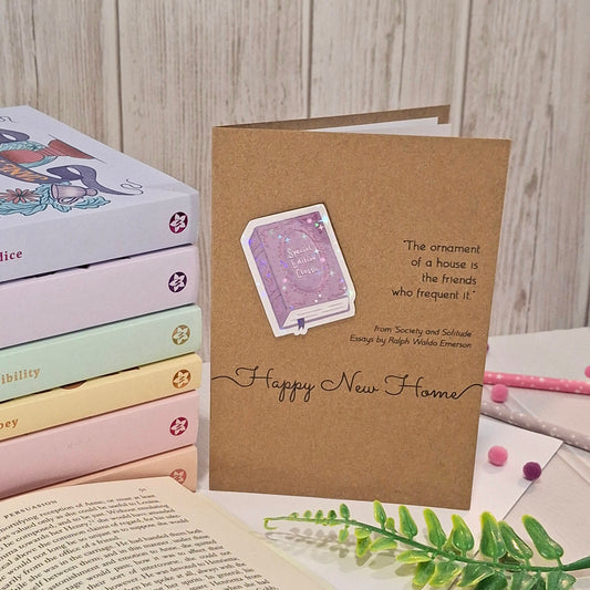 A brown kraft greeting card styled on a tabletop with books, pastel pens, and greenery. The card features a raised holographic embellishment of a purple special edition classic book, the word Happy New Home, and a quote from Ralph Waldo Emerson.