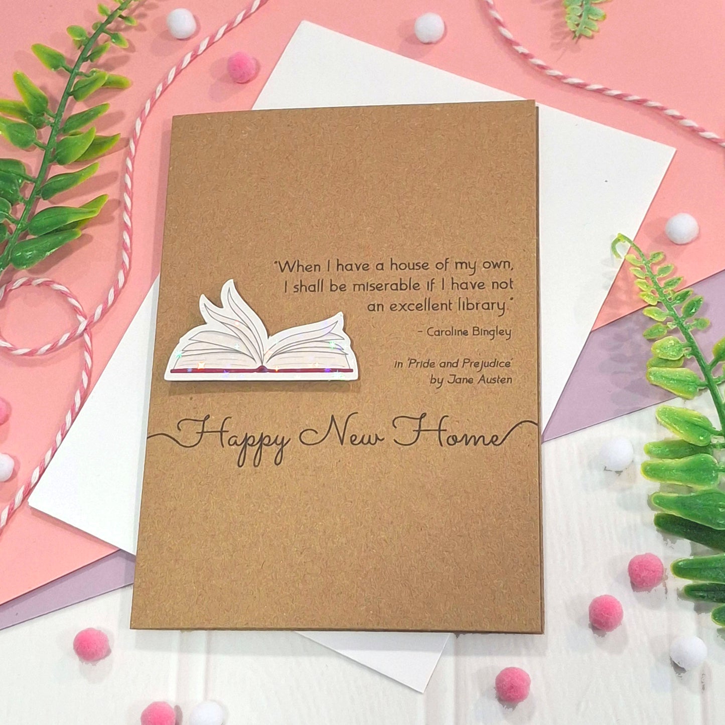 A brown kraft new home card with illustrated book pages in a raised, holographic embellishment, the words Happy New Home, and a quote from Jane Austen's Pride and Prejudice, styled on a pink and white surface with decorative greenery and pom-poms.