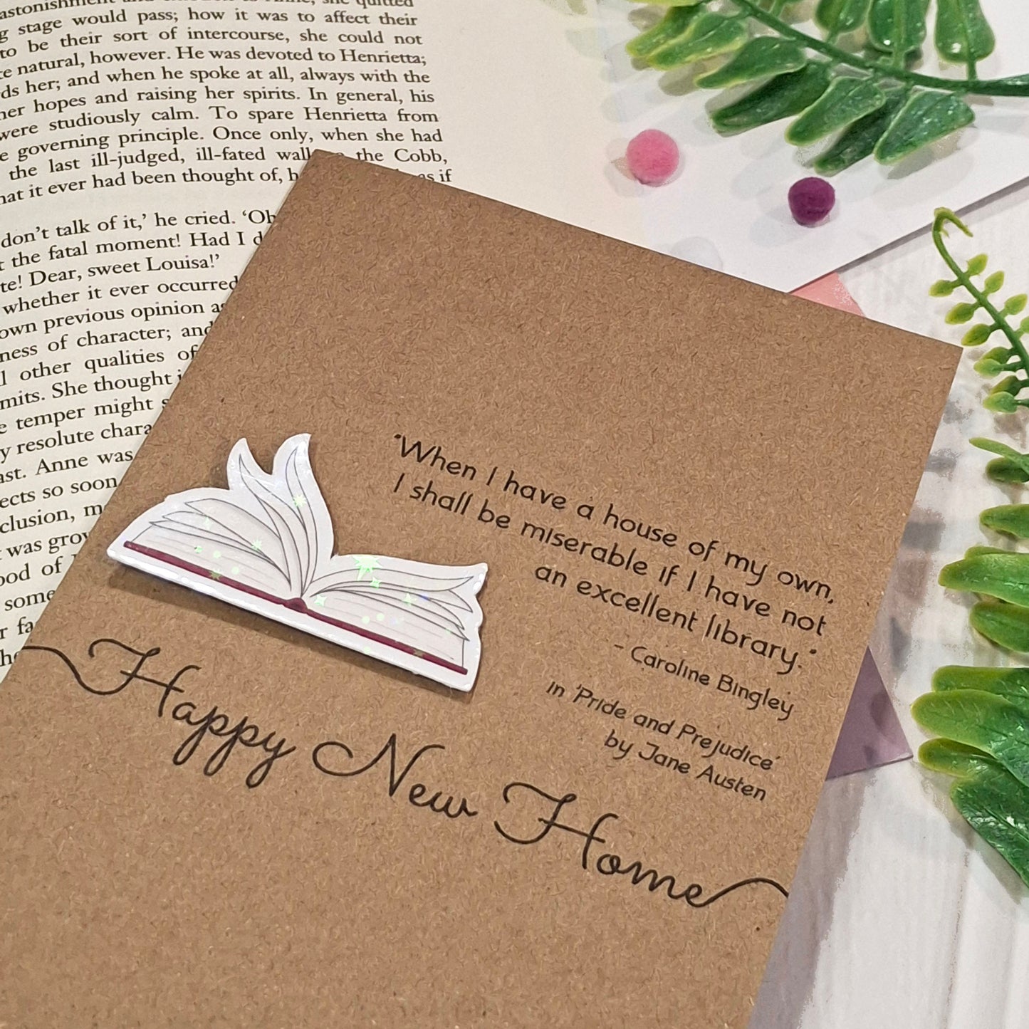 A kraft card styled on top of an open book with small decorative flowers and green leaves. The card features a raised holographic embellishment of an open book with gently fanned pages, the words Happy New Home, and a quote from the book Pride and Prejudice by Jane Austen.