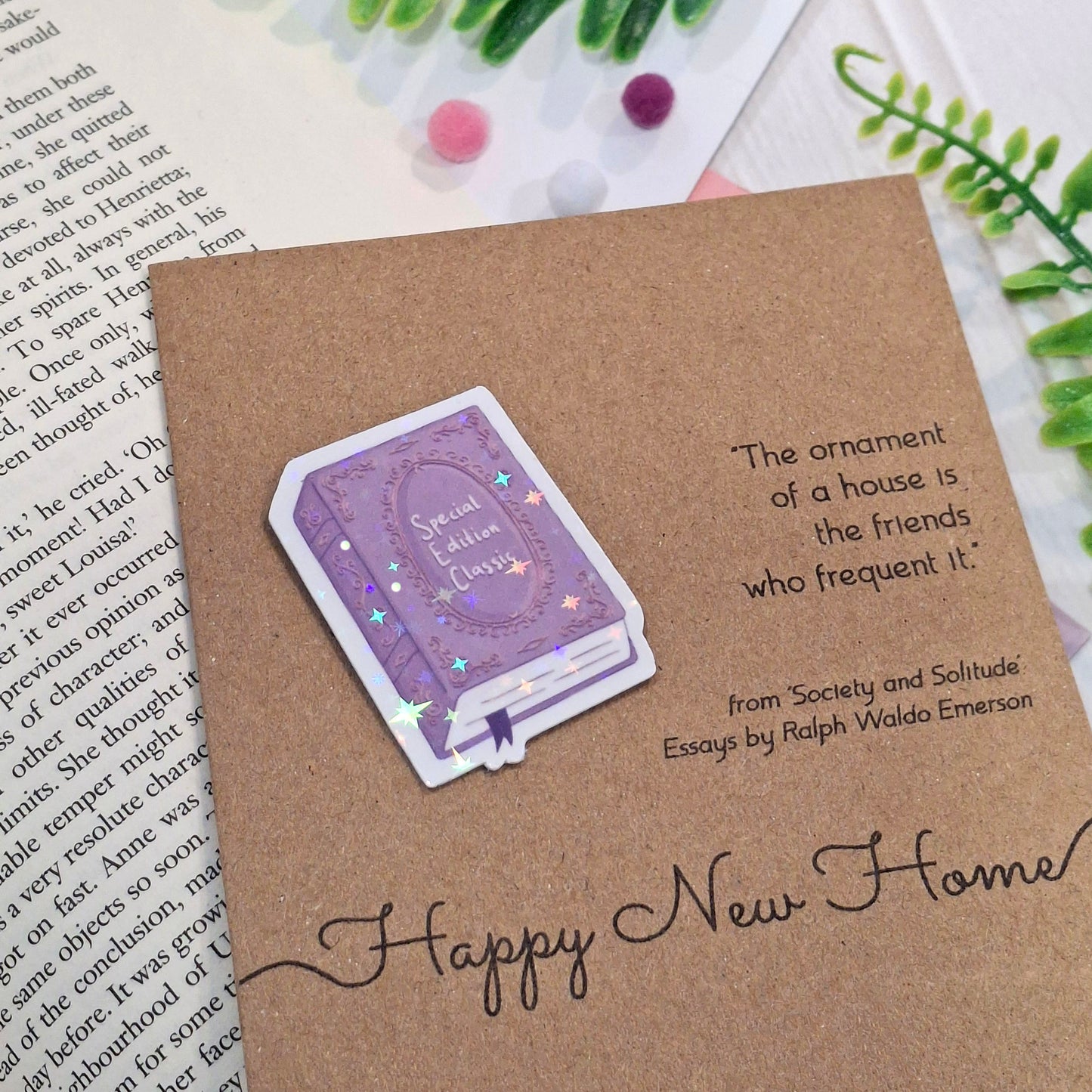 A kraft paper card styled on top of an open book with small decorative pom-poms and greenery. The card features a raised holographic embellishment of a purple special edition classic book, the word Happy New Home, and a quote from Ralph Waldo Emerson.