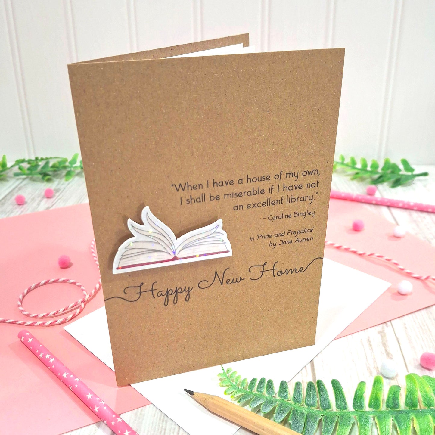 A brown kraft new home card with illustrated book pages in a raised, holographic embellishment, the words Happy New Home, and a quote from Jane Austen's Pride and Prejudice. The card is displayed on a styled flat-lay surface with pink paper, a pencil, and green leaves.