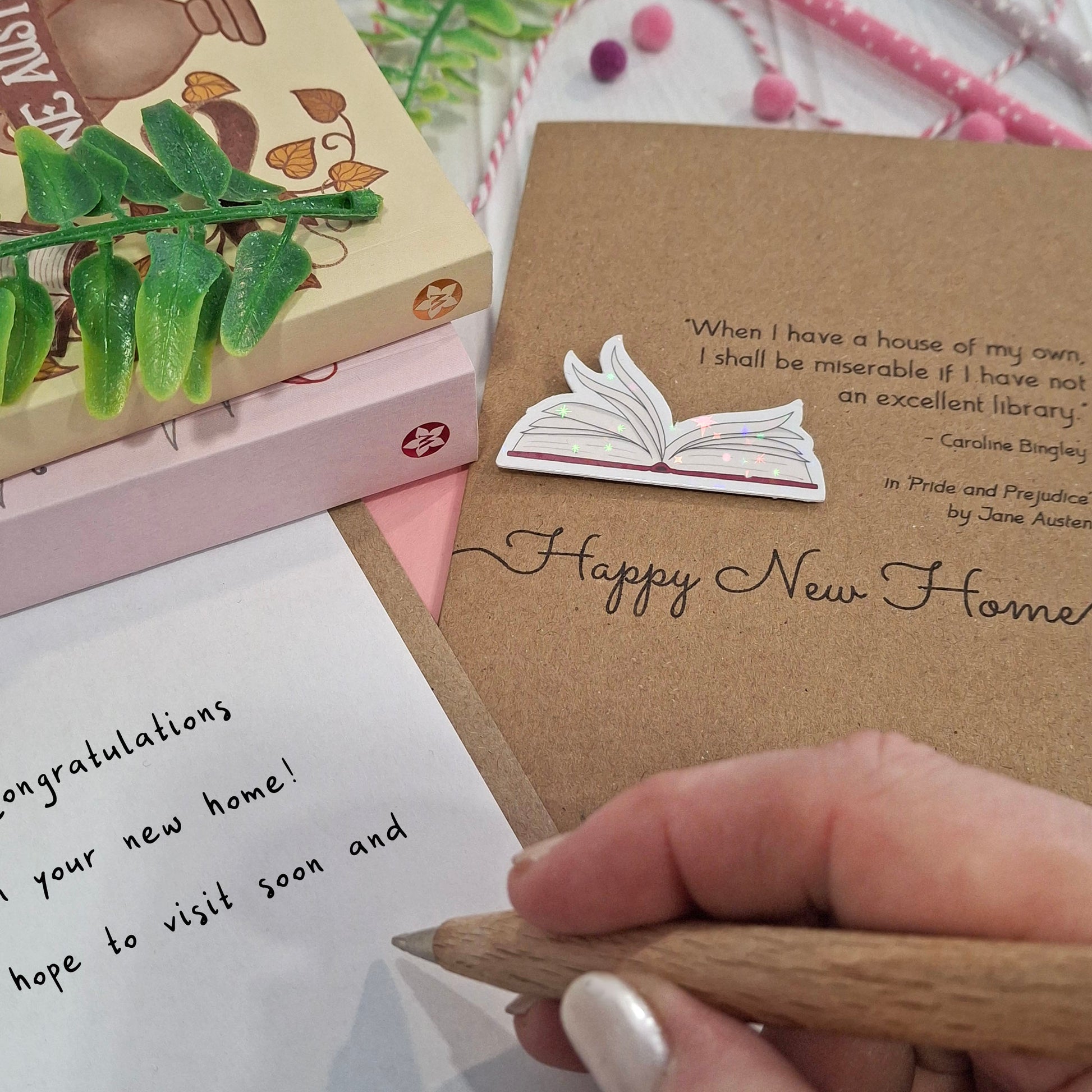 A close up of a hand holding a wooden pen, writing a congratulations message inside a card. The card features a raised holographic embellishment of an open book with gently fanned pages, the words Happy New Home, and a quote from the book Pride and Prejudice by Jane Austen.