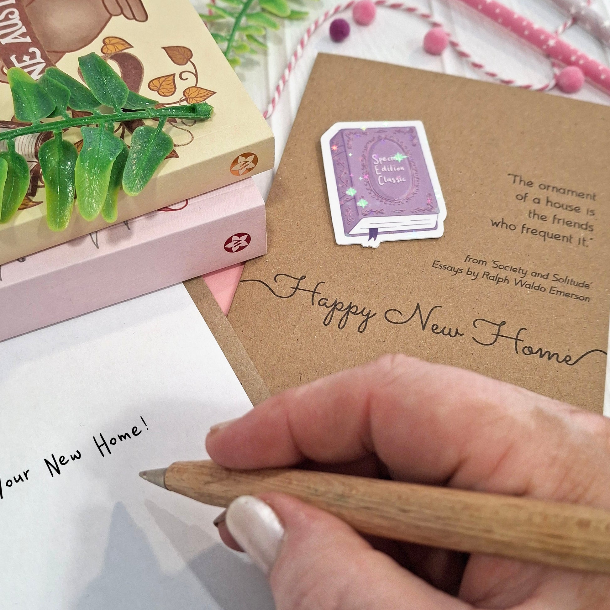 A close-up of a hand holding a wooden pencil writing a message inside a card. The card features a raised holographic embellishment of a purple special edition classic book, the word Happy New Home, and a quote from Ralph Waldo Emerson.