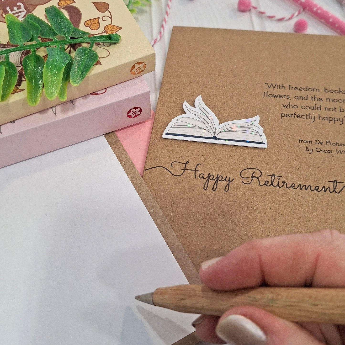 A close up of a hand holding a wooden pen, and writing inside a card. The card features a raised holographic embellishment of an open book with gently fanned pages, the words Happy Retirement, and a quote by Oscar Wilde.