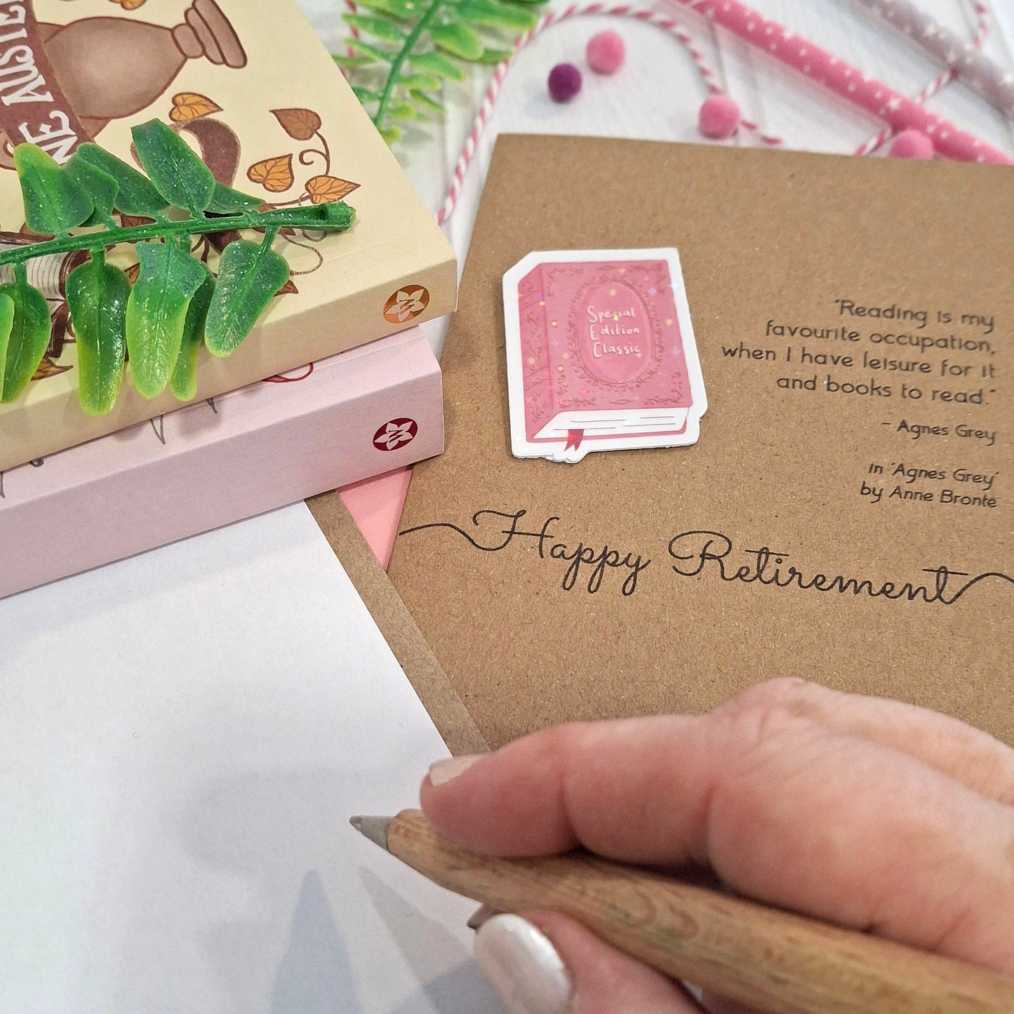 A close-up of a hand holding a wooden pen and writing inside a card. The card features a raised holographic embellishment of a pink special edition classic book, the words Happy Retirement, and a quote from Agnes Grey by Anne Bronte.