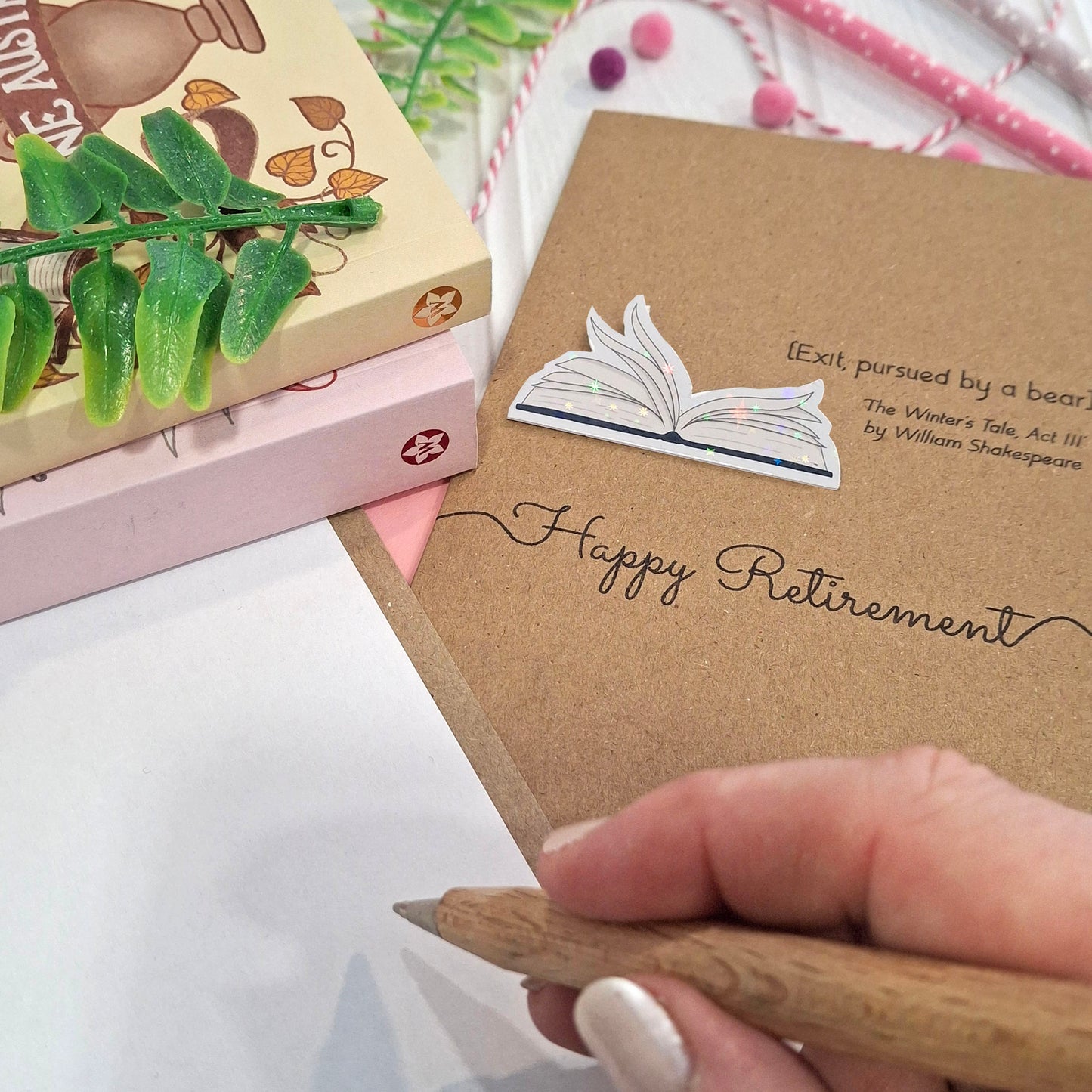 A close up of a hand holding a wooden pen, and writing inside a card. The card features a raised holographic embellishment of an open book with gently fanned pages, the words Happy Retirement, and a quote from The Winter's Tale by William Shakespeare.