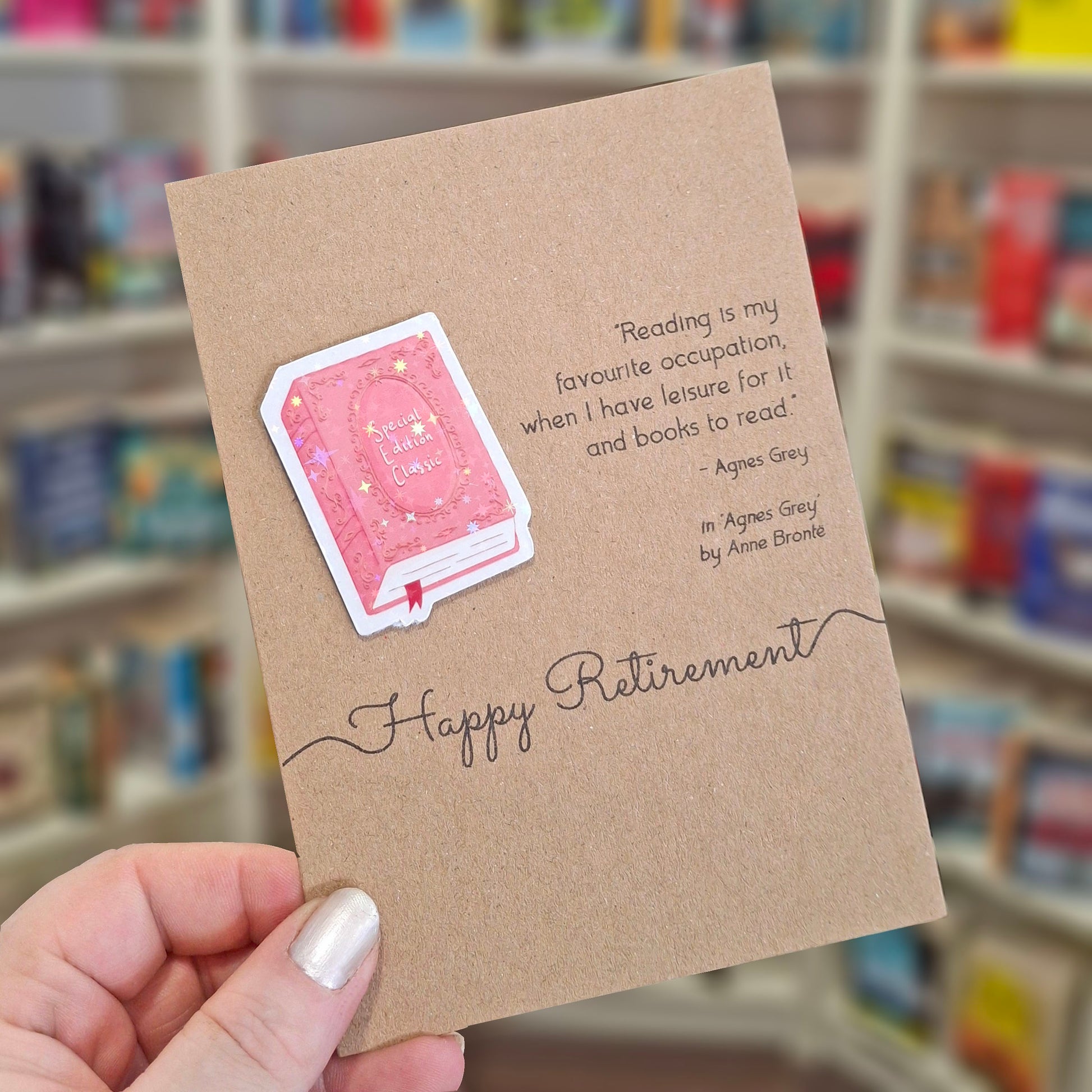 A hand holds a brown kraft happy retirement card featuring a raised holographic embellishment of a pink special edition classic book, and a printed quote by Anne Bronte. The background shows a blurred bookshop interior.