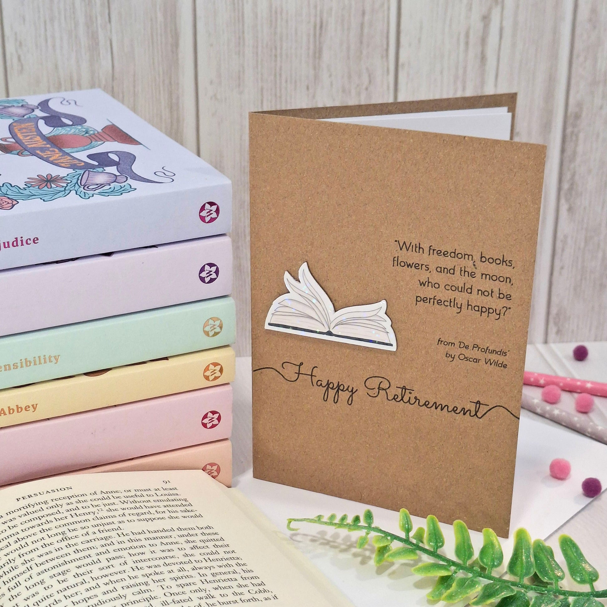 A brown kraft retirement card styled on a tabletop with books and greenery. The card features a raised holographic embellishment of an open book with gently fanned pages, the words Happy Retirement, and a quote by Oscar Wilde.