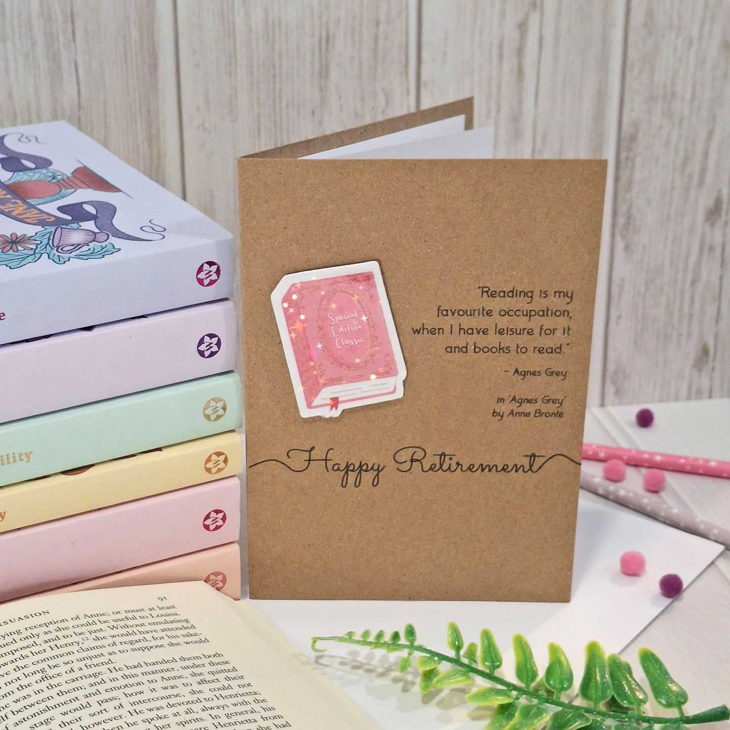 A retirement card made from brown kraft card styled on a light wood background with books, an open book, and pens. The card features a raised holographic embellishment of a pink special edition classic book, the words Happy Retirement, and a quote from Agnes Grey by Anne Bronte.