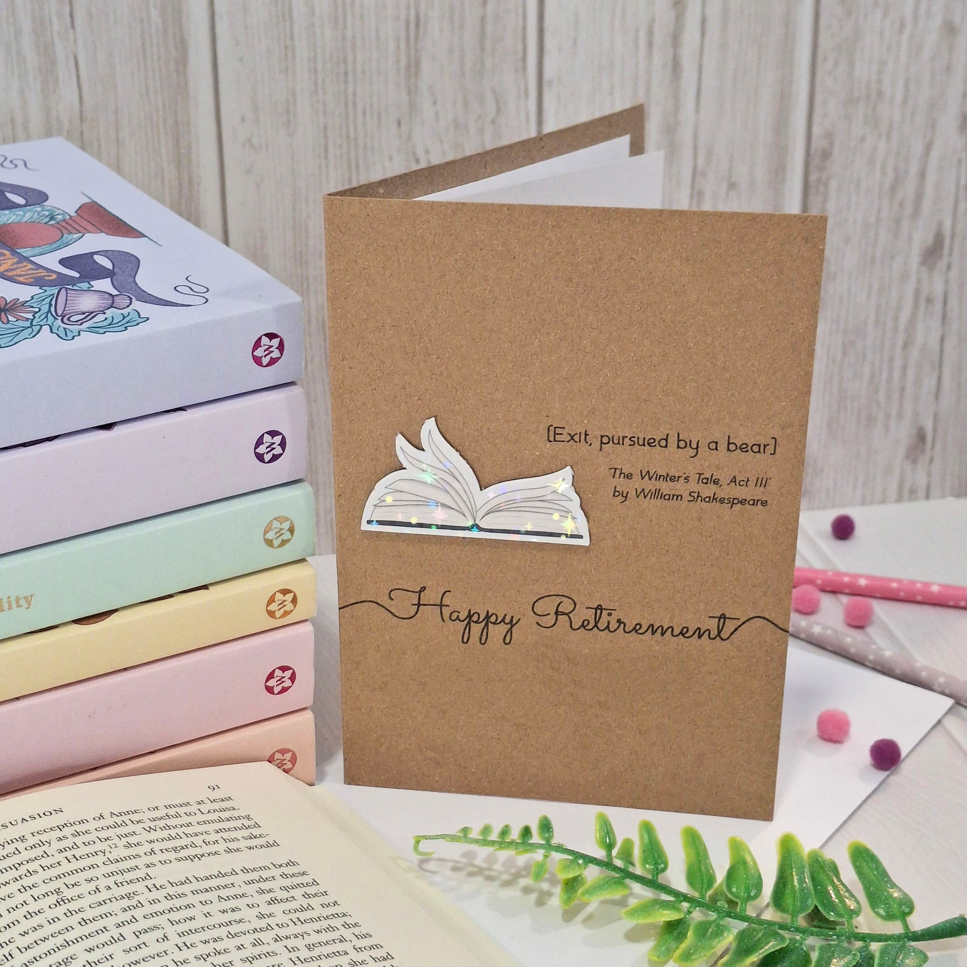 A brown kraft paper retirement card styled with a pastel book stack, and greenery. The card features a raised holographic embellishment of an open book with gently fanned pages, the words Happy Retirement, and a quote from The Winter's Tale by William Shakespeare.