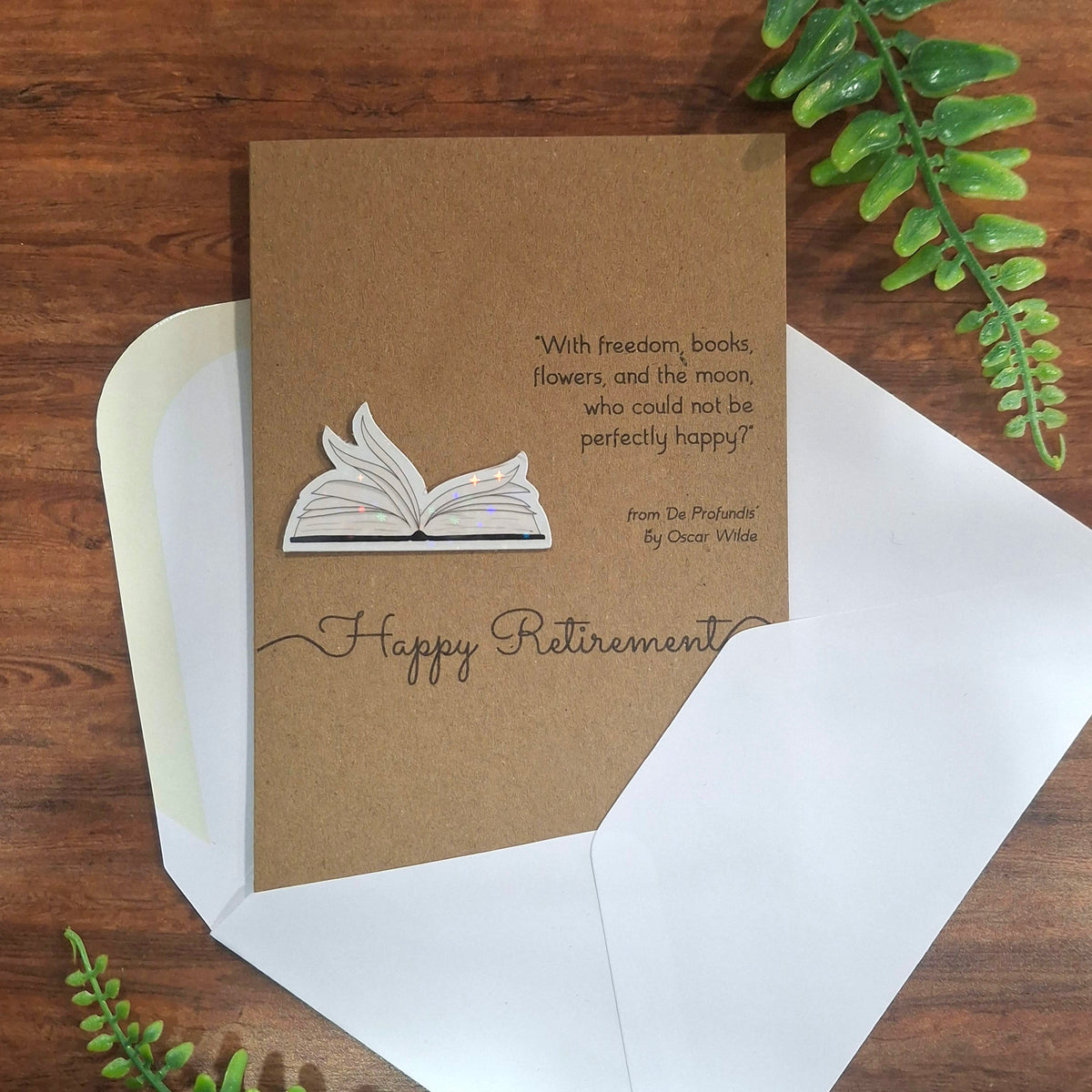 A Happy Retirement greeting card and white envelope laid on a wooden surface with green leaves all around it. The card is made from brown kraft card and features a raised 3D holographic embellishment of open fluttering book pages, and a quote from De Profundis by Oscar Wilde.
