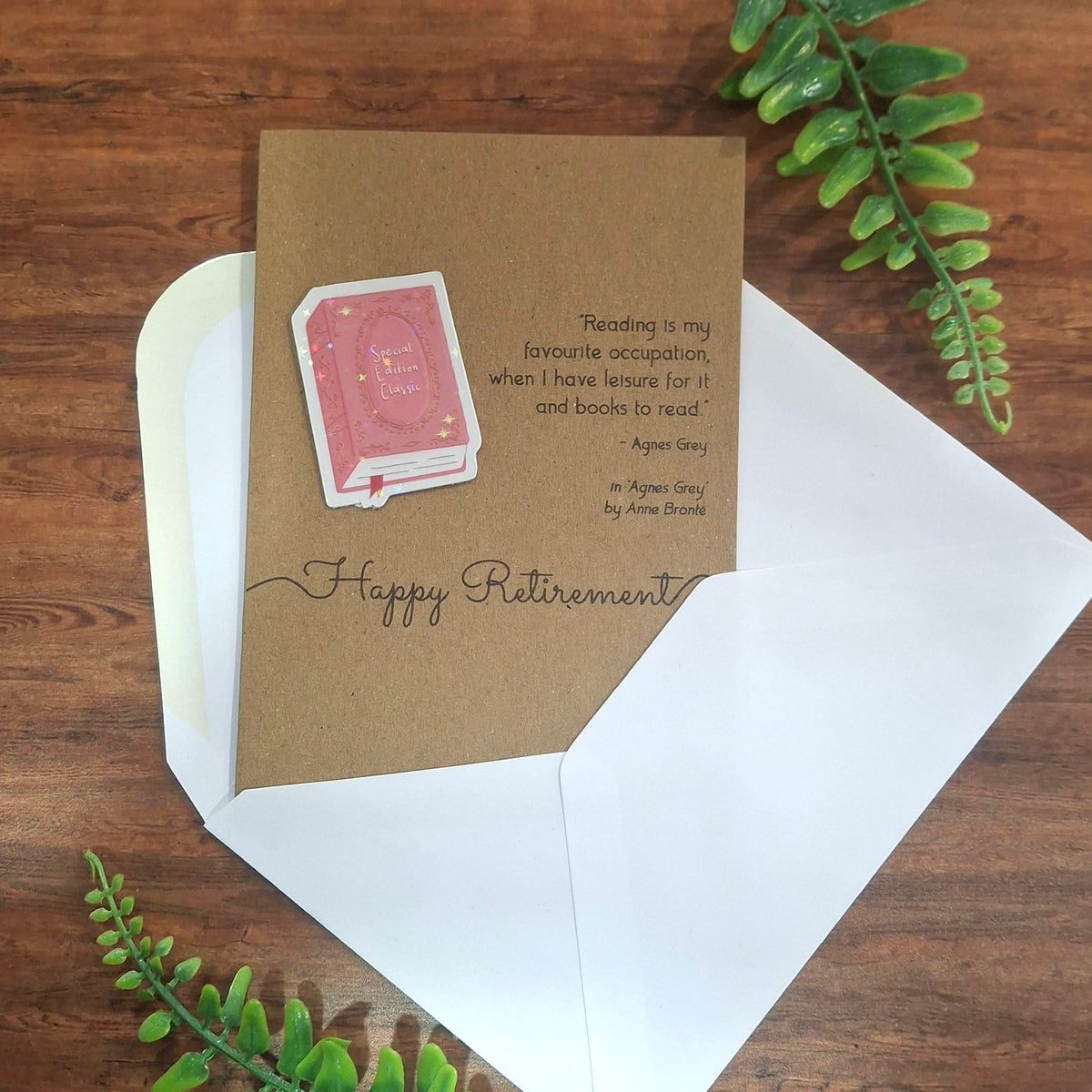 A Happy Retirement greeting card and white envelope laid on a wooden surface with green leaves around it. The card is made from brown kraft card and features with a raised 3D holographic pink book cover embellishment, and a quote from Agnes Grey by Anne Bronte. 