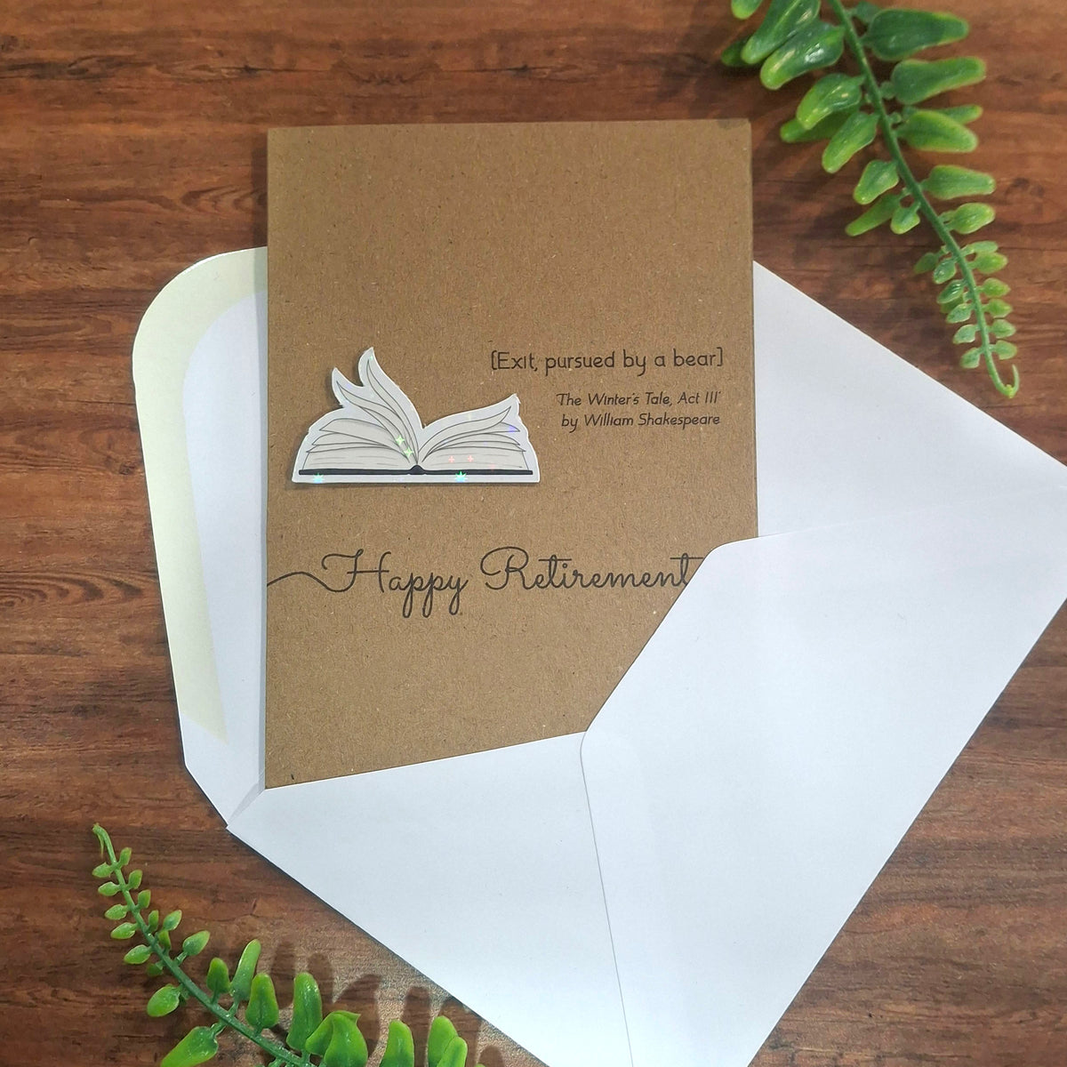 A Happy Retirement greeting card and white envelope laid on a wooden surface with green leaves all around it. The card is made from brown kraft card and features a raised 3D holographic embellishment of open fluttering book pages, and a quote from The Winter.s Tale Act III by William Shakespeare.