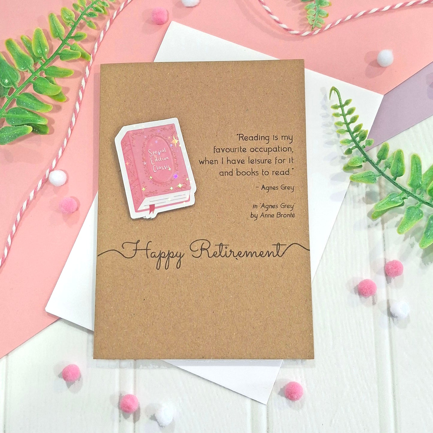 A kraft retirement card with a raised, holographic, illustrated story book embellishment in pink, a quote from Agnes Grey by Anne Bronte, and the words 'Happy Retirement' in elegant script, all styled on a pink and white flat-lay background with decorative greenery and pom-poms.