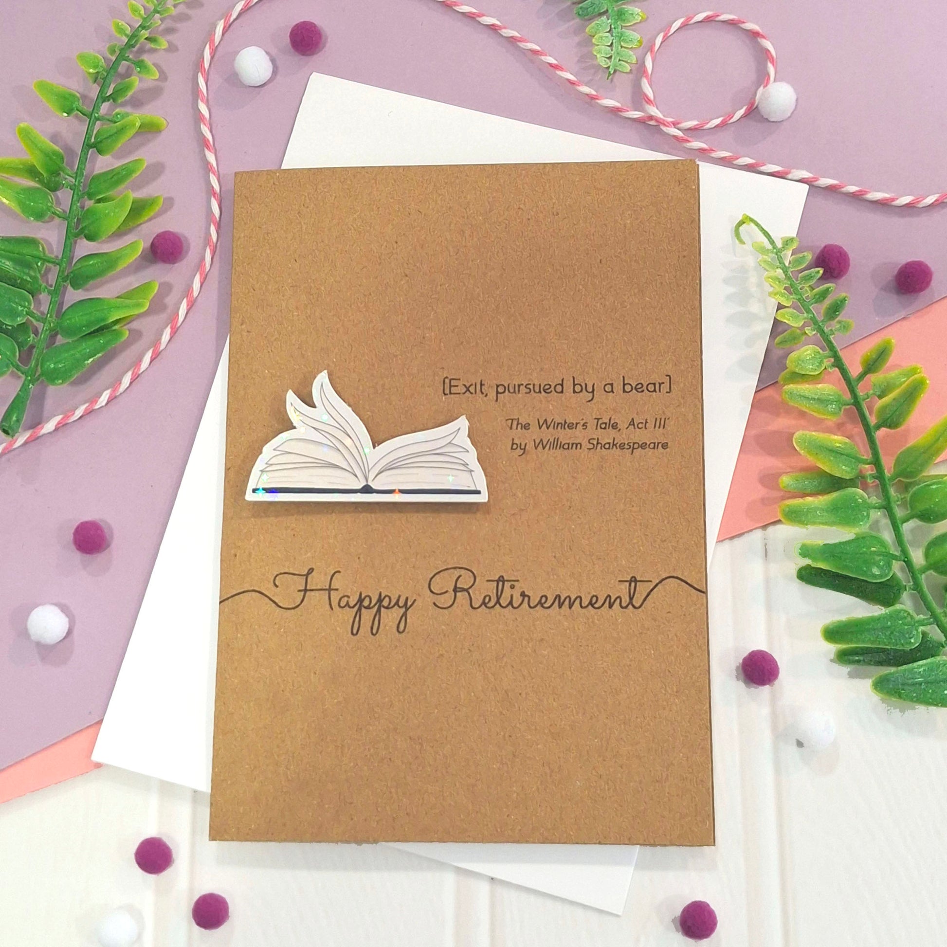 A brown kraft retirement card with illustrated book pages in a raised, holographic embellishment, the words Happy Retirement, and a quote from William Shakespeare's The Winter's Tale Act III, all styled on a pastel flat-lay with green leaves, pom-poms, and ribbon.
