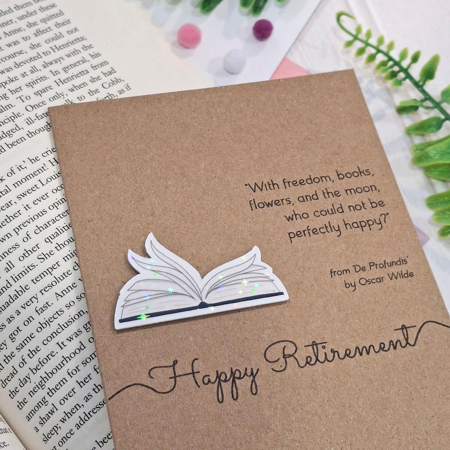 A brown kraft retirement card styled on an open book with green leaves. The card features a raised holographic embellishment of an open book with gently fanned pages, the words Happy Retirement, and a quote by Oscar Wilde.