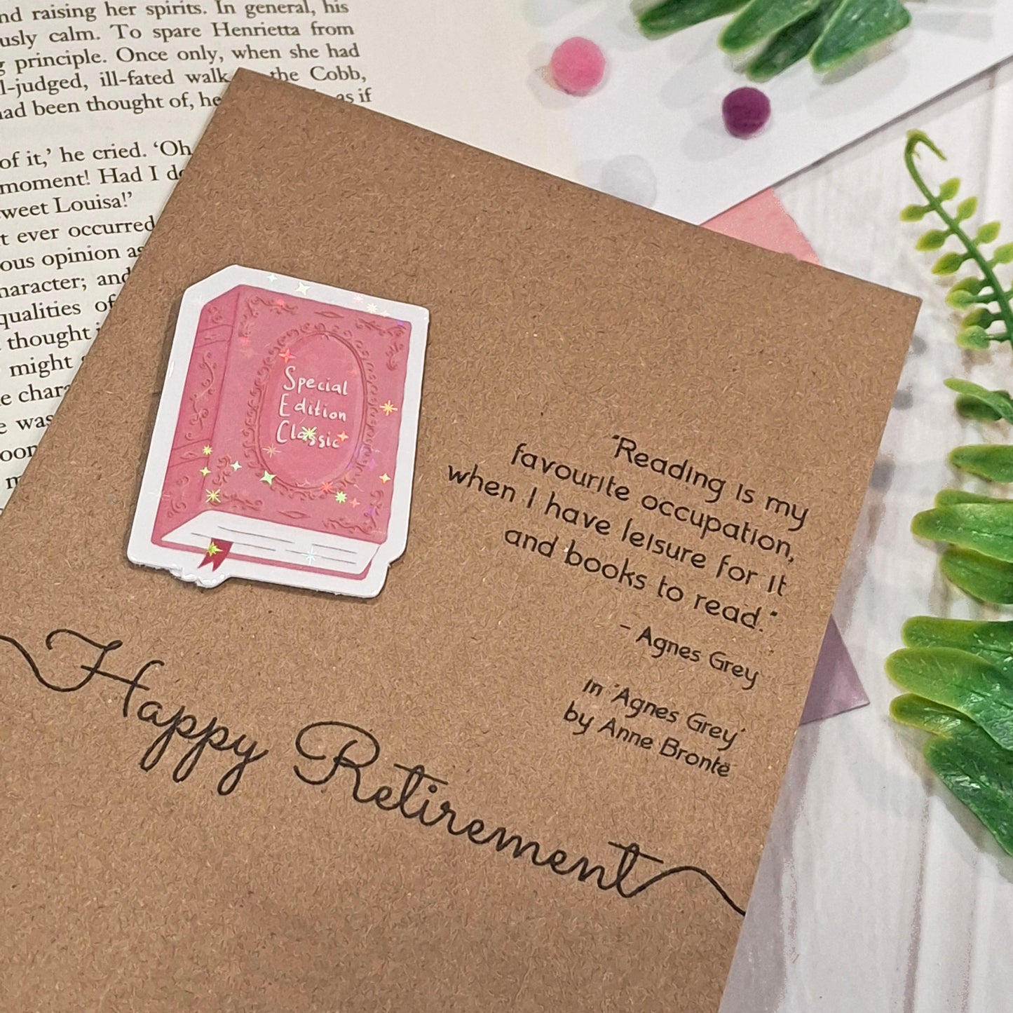 A retirement card made from brown kraft card displayed on a light wood surface with decorative green leaves and a book page in the background. The card features a raised holographic embellishment of a pink special edition classic book, the words Happy Retirement, and a quote from Agnes Grey by Anne Bronte.