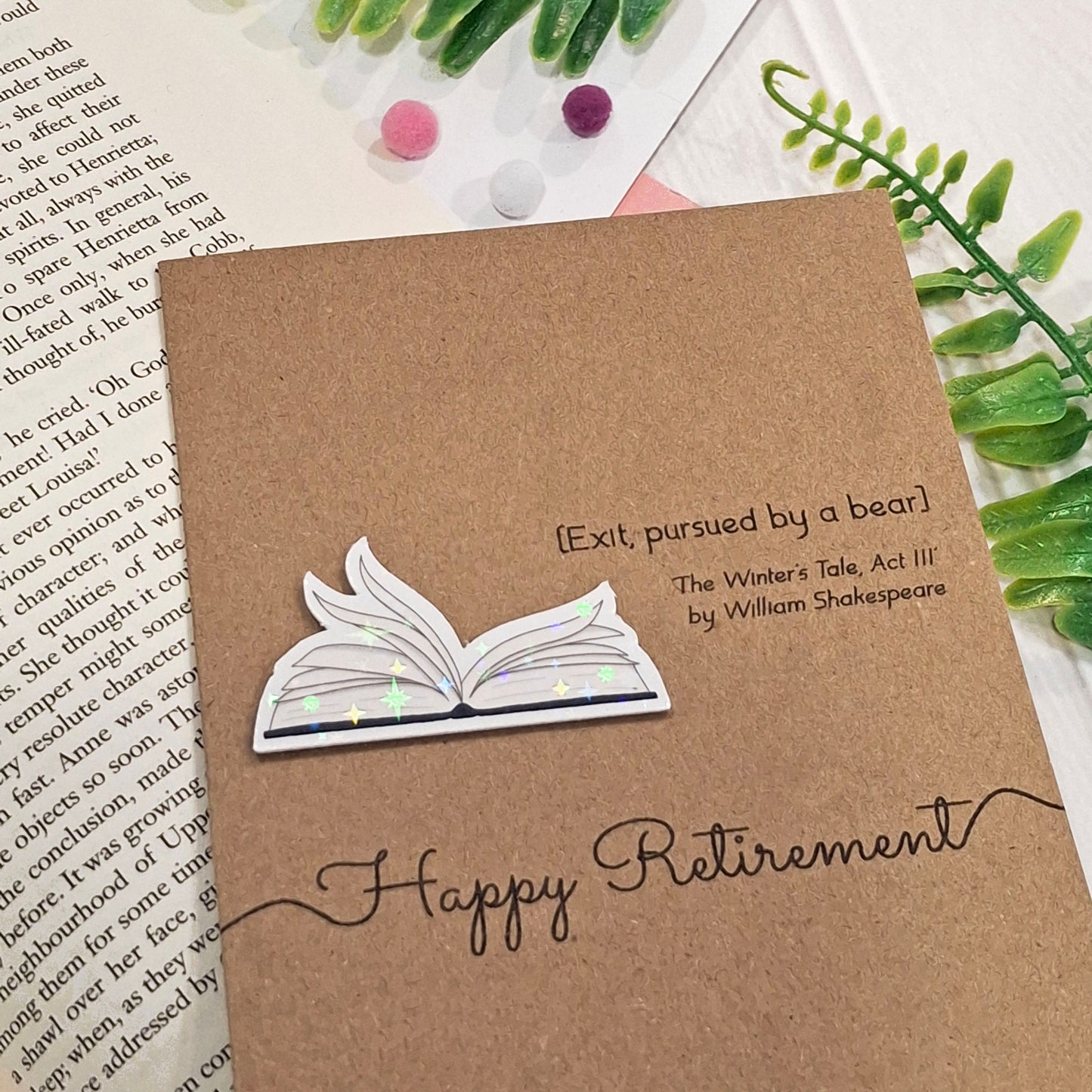 A brown kraft retirement card placed on an open book with decorative green leaves and pom-poms. The card features a raised holographic embellishment of an open book with gently fanned pages, the words Happy Retirement, and a quote from The Winter's Tale by William Shakespeare.