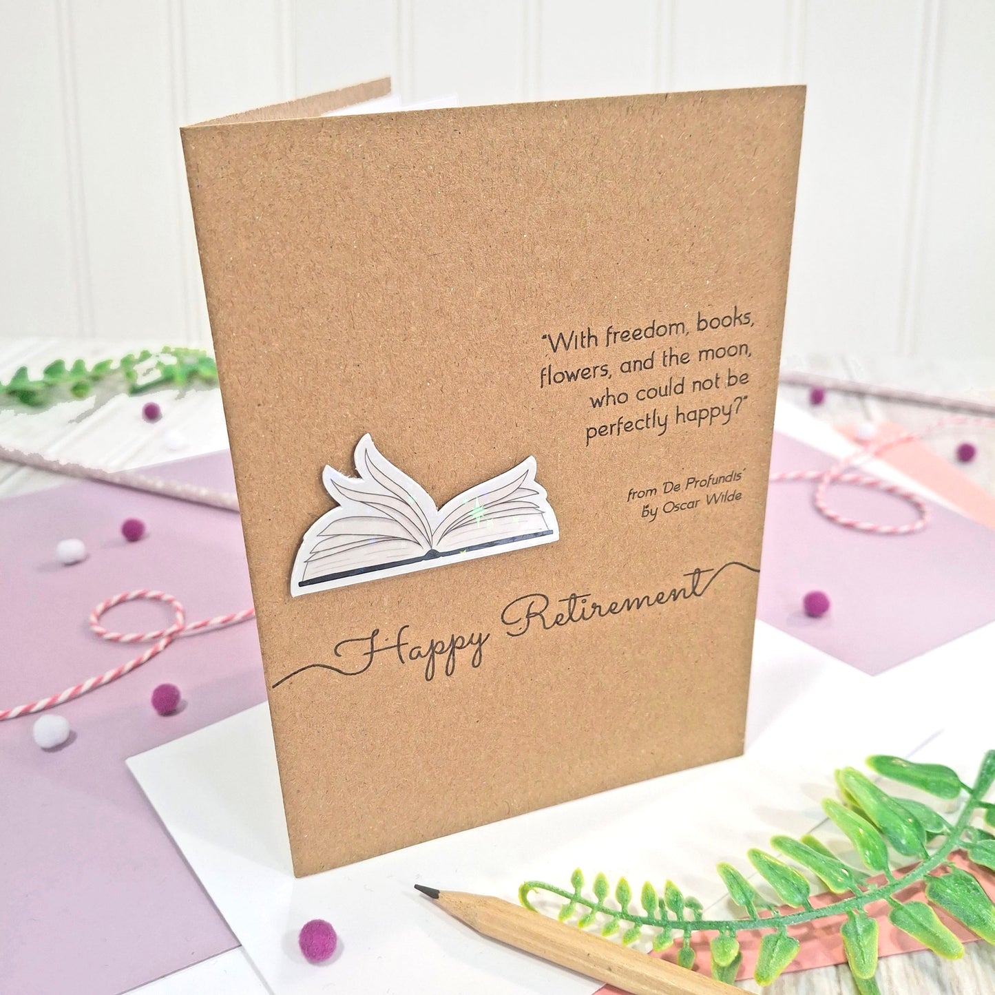 A handmade retirement card made from brown kraft card with illustrated book pages in a raised, holographic embellishment, the text 'Happy Retirement' and a quote by Oscar Wilde. The card is displayed standing up on a styled flat-lay surface with a pencil, greenery, and decorative ribbons.