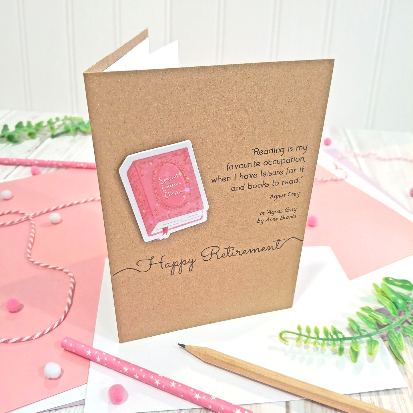 A brown kraft retirement card standing upright, decorated with a raised, holographic, illustrated story book embellishment in pink, a quote from Agnes Grey by Anne Bronte, and 'Happy Retirement' in black script. The card is styled on a white surface with pink paper, a wooden pencil, and greenery.